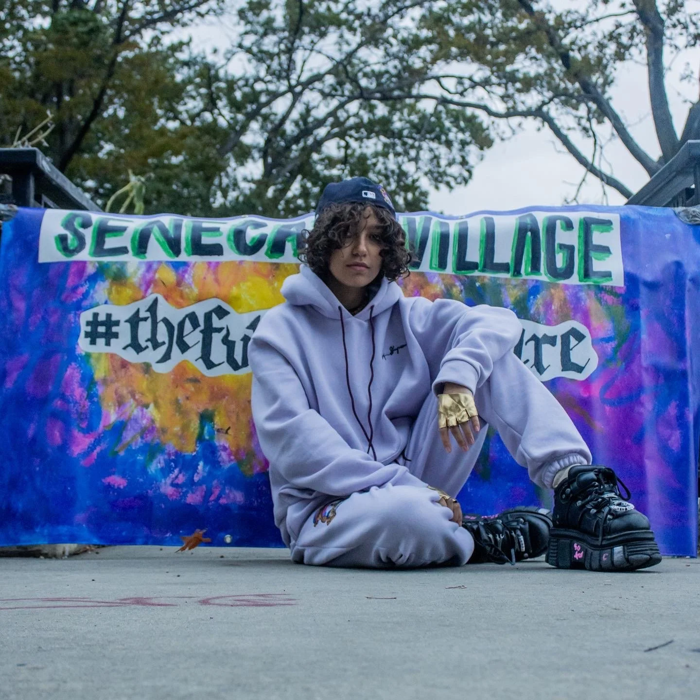 A person in a gray hoodie and sweatpants with blond hair and golden gloves sitting on the ground in front of a colorful banner that reads 'SENECA VILLAGE' and includes hashtags and other text, with trees and cloudy sky in the background.