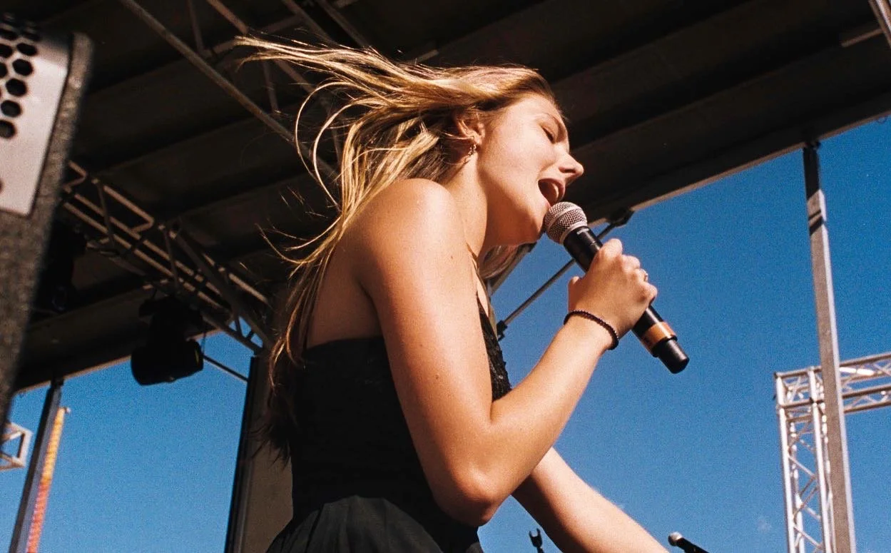 Young woman singing into a microphone on an outdoor stage with blue sky and metal scaffolding in the background.
