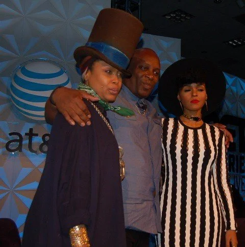 Three people posing for a photo at an event, with two women dressed stylishly and a man in the middle. The woman on the left wears a large brown hat with a blue band and a dark outfit. The woman on the right wears a wide-brimmed black hat and a black