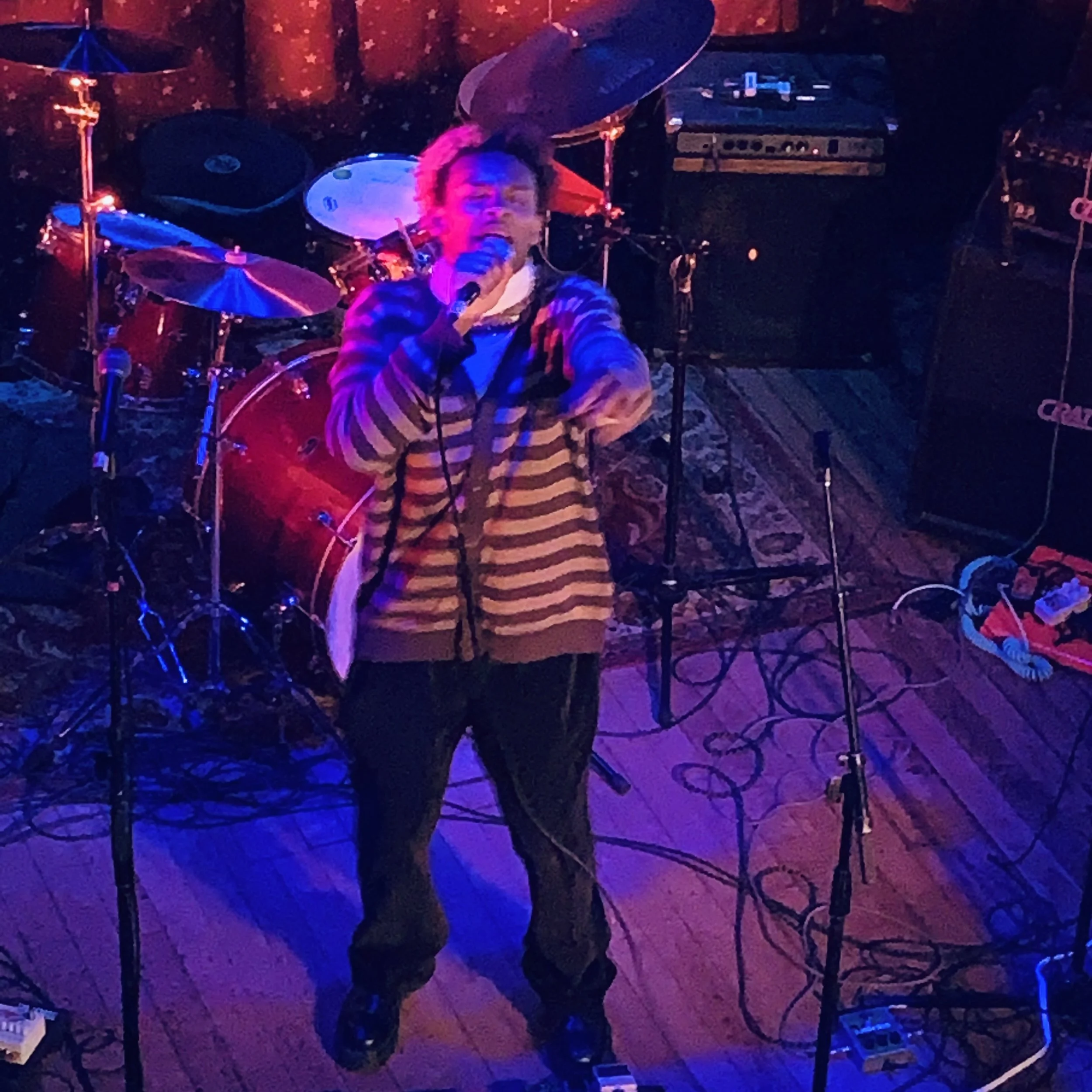 A person singing into a microphone on stage with musical instruments and equipment behind them, including a drum set and guitar amps, under colorful stage lighting.