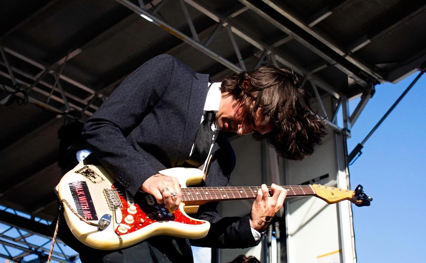 Musician playing electric guitar on stage during outdoor concert, wearing a dark suit and white shirt, with blue sky background.