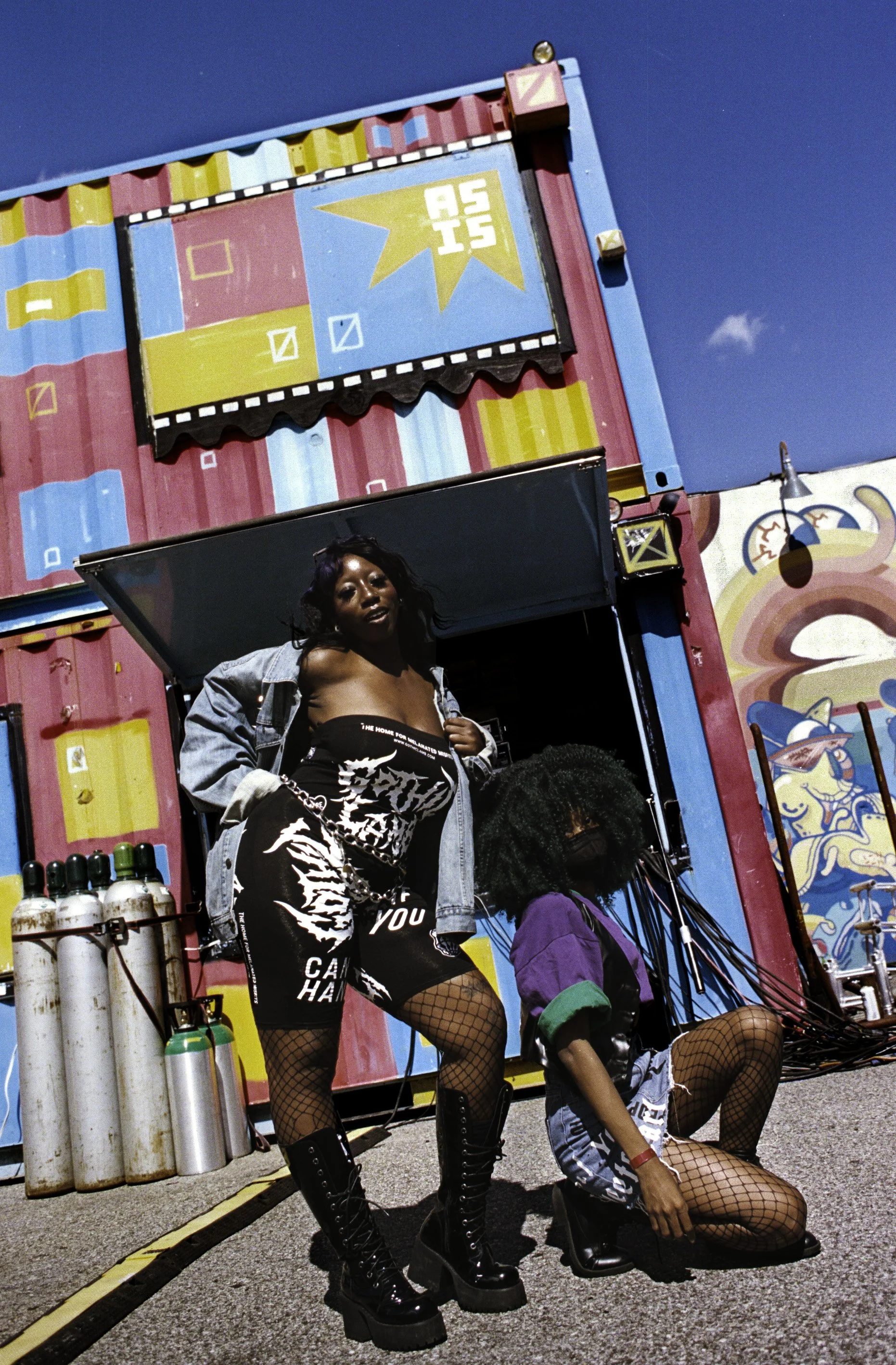 Two women with curly hair and fishnet stockings pose in front of a colorful amusement park ride with bright pink, blue, and yellow paint and graphic art.