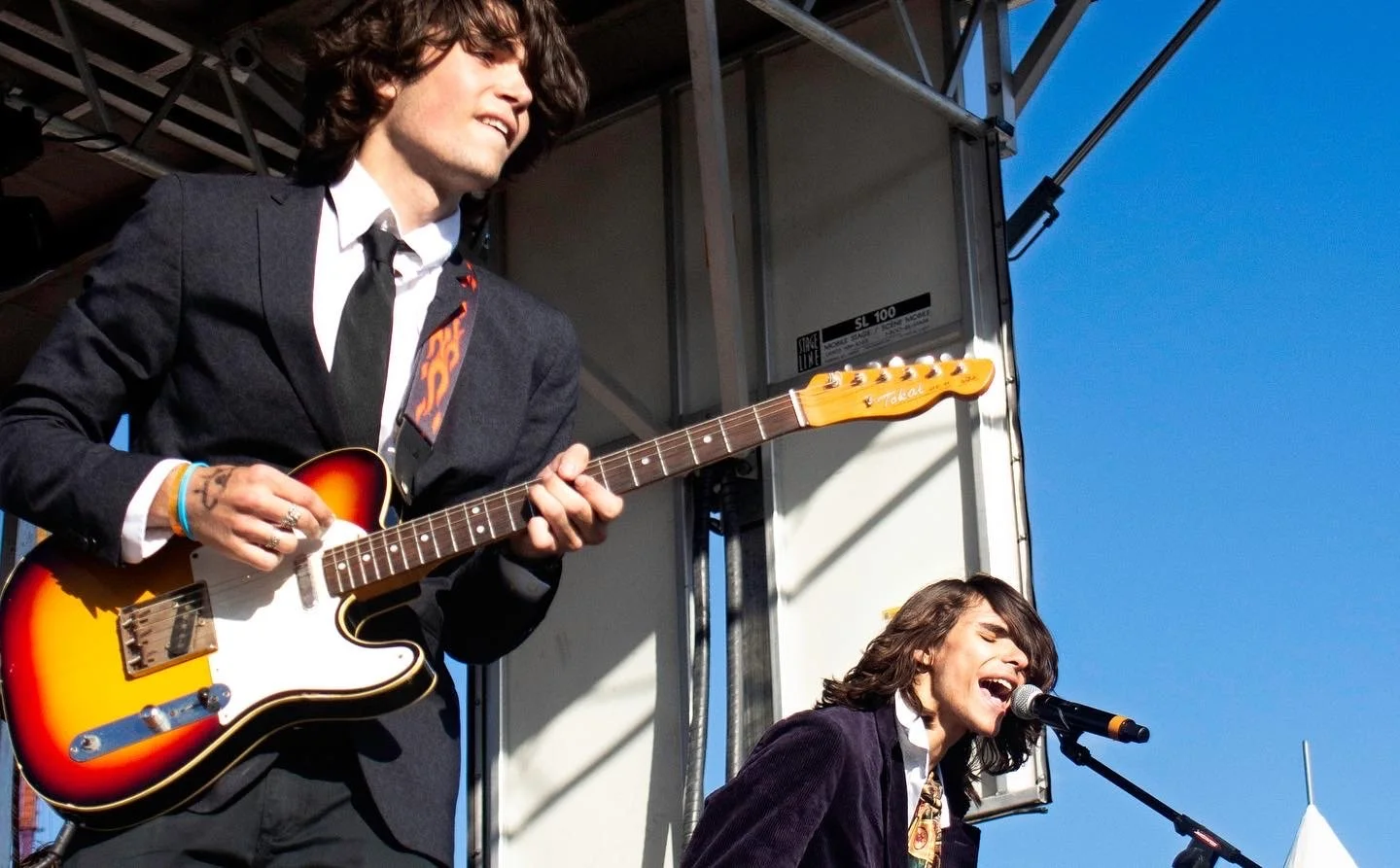 Two musicians performing on a stage outdoors. The one on the left is playing an electric guitar; he has dark hair, is dressed in a suit, and is looking to the right. The one on the right is singing into a microphone with a joyful expression, wearing 