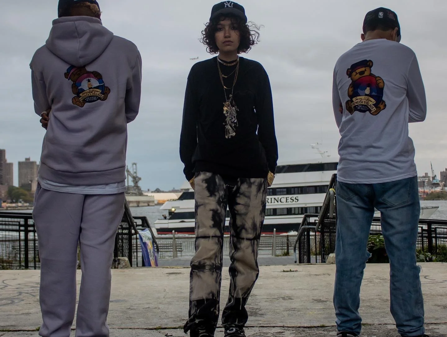 Three young people standing near a waterfront, with a large yacht labeled 'PRINCESS' in the background and city buildings beyond.