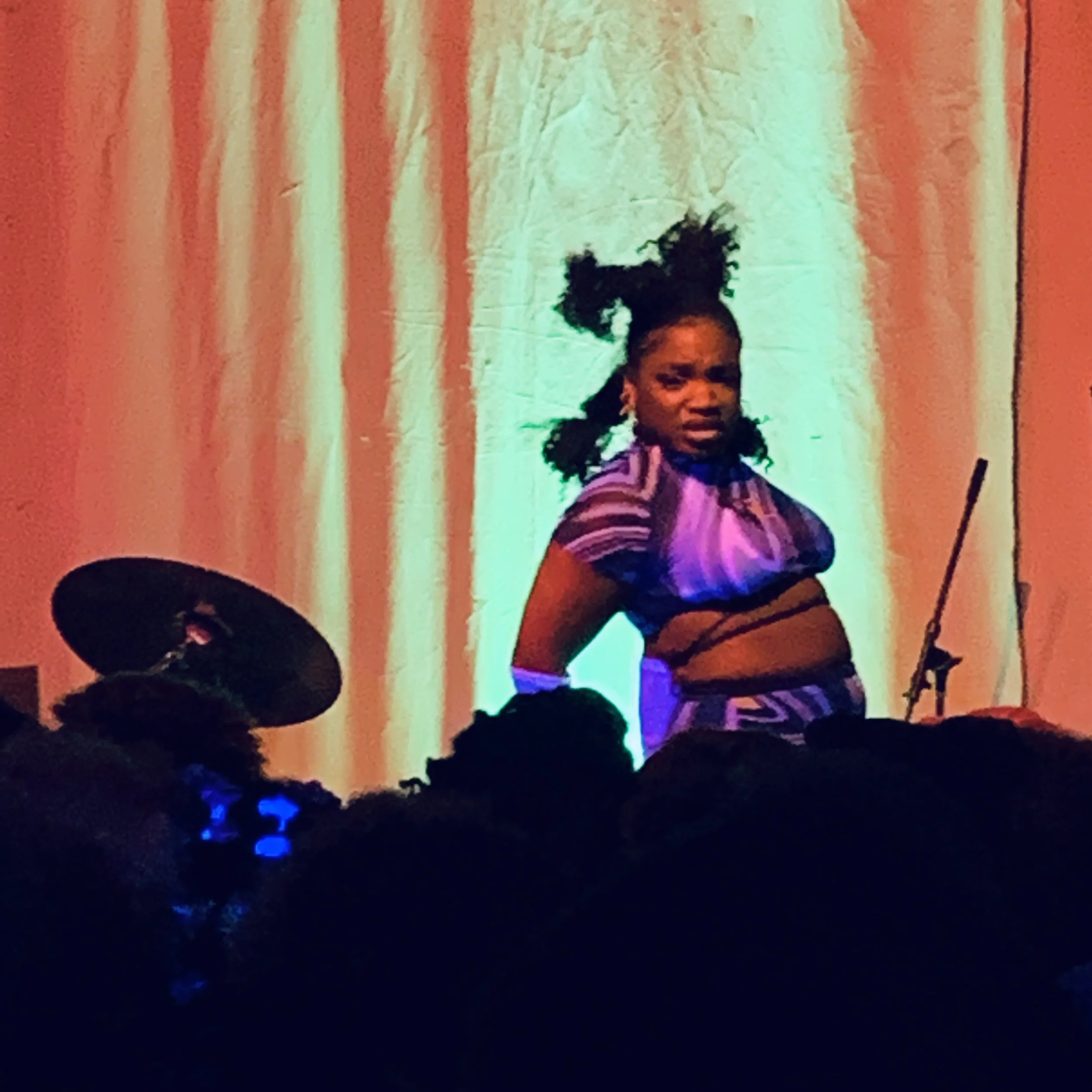 Young girl with curly hair in pigtails standing on a stage with a backdrop, wearing a colorful outfit, and looking determined. A drum kit is partially visible to the left, and the audience is in silhouette at the bottom of the image.