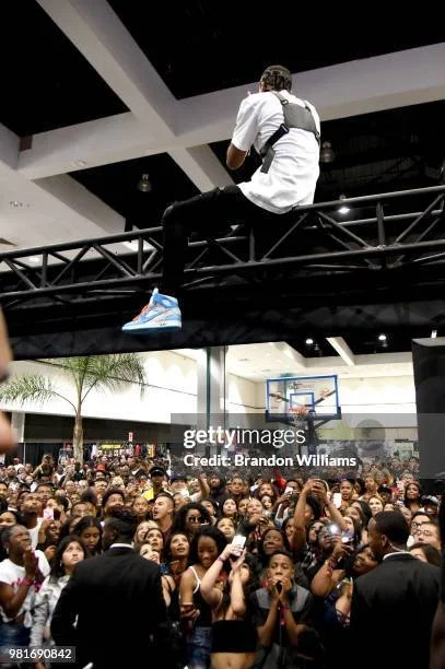 A man sitting on a black metal railing above a crowd of people, many holding up cameras or phones, inside a large indoor space with a basketball hoop in the background.