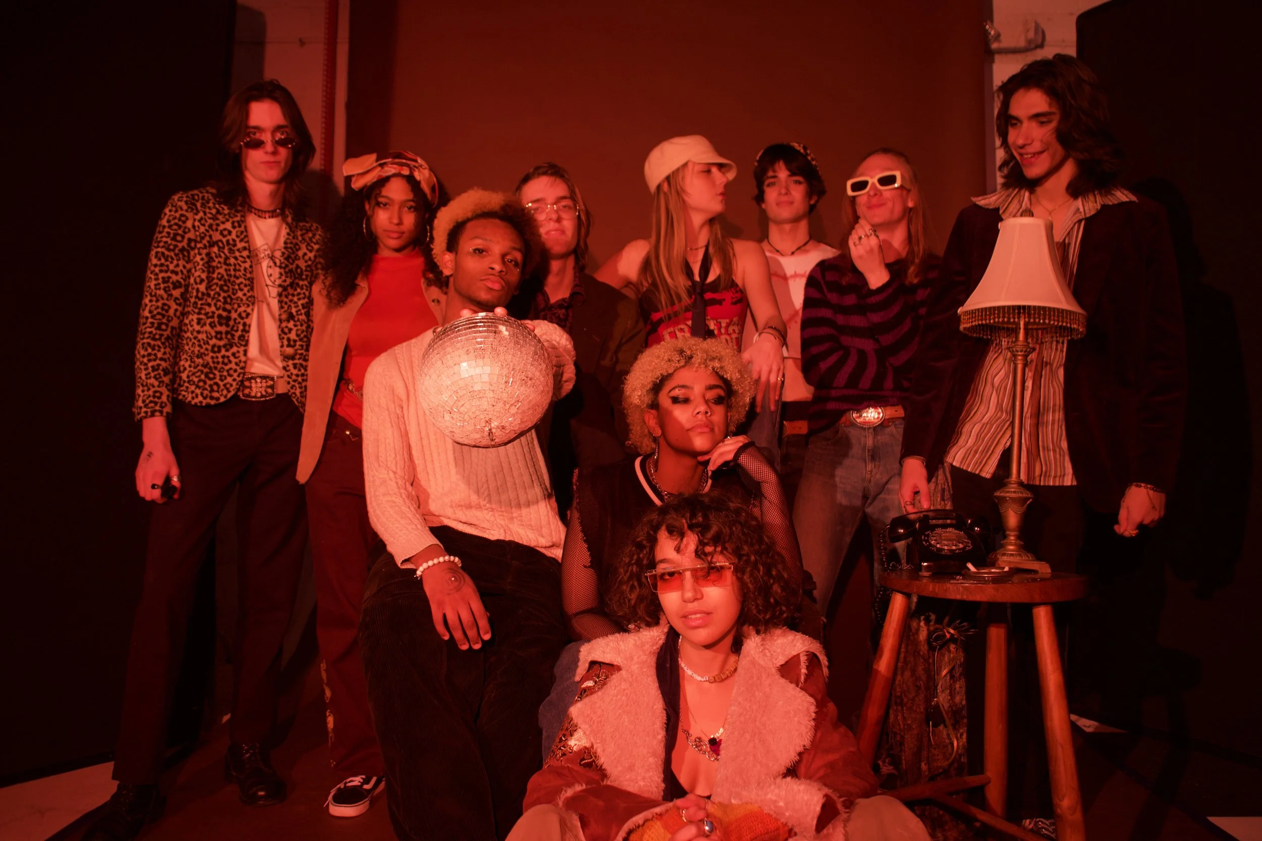 Group of diverse young people posing on stage with red lighting, some wearing vintage and retro outfits, standing and sitting around a small wooden table with a lamp and a vintage rotary phone.