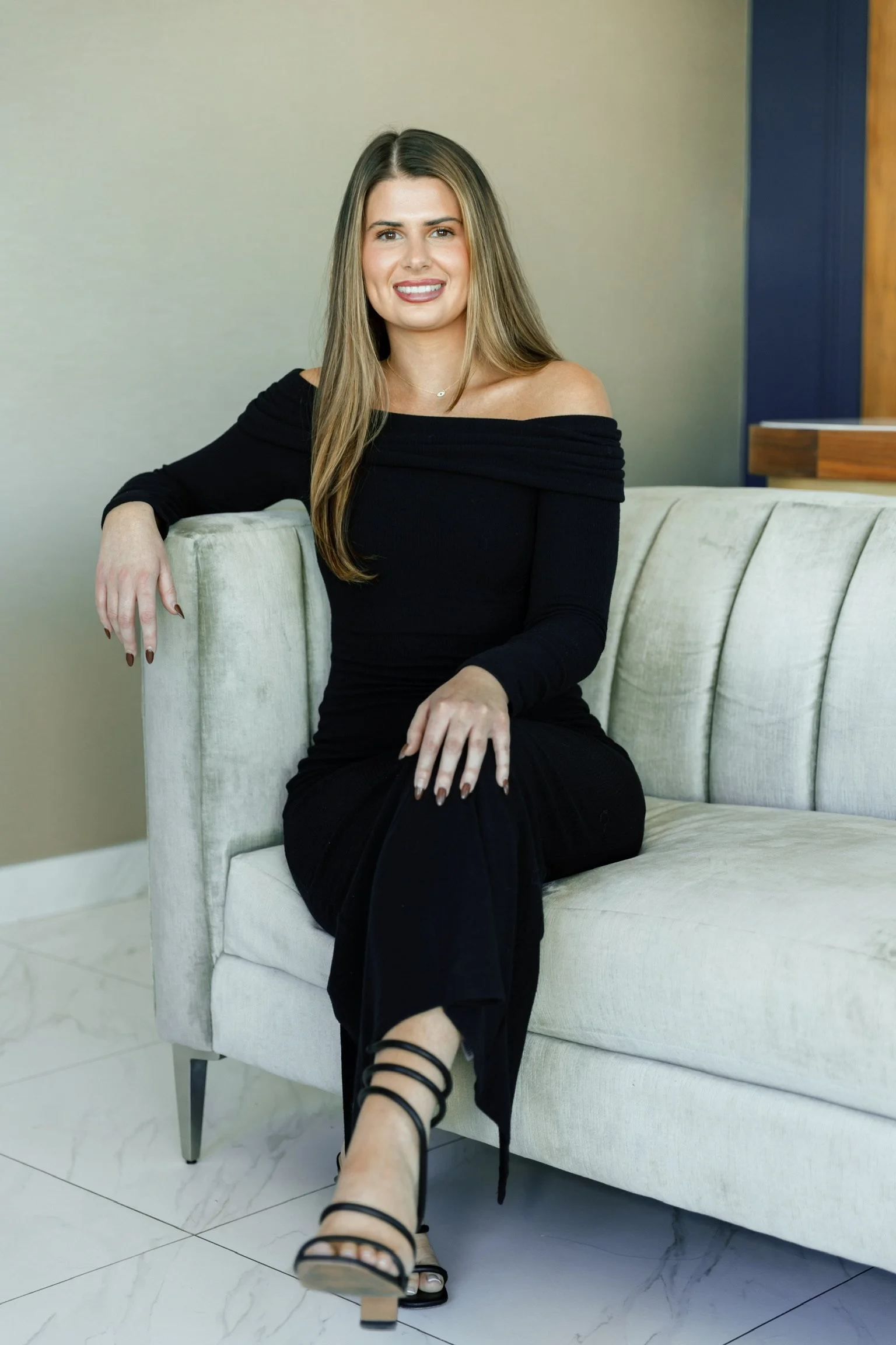 A woman with long blonde hair and a black off-shoulder dress sitting on a white sofa, smiling at the camera. She is in a modern room with marble flooring and a blue accent wall.