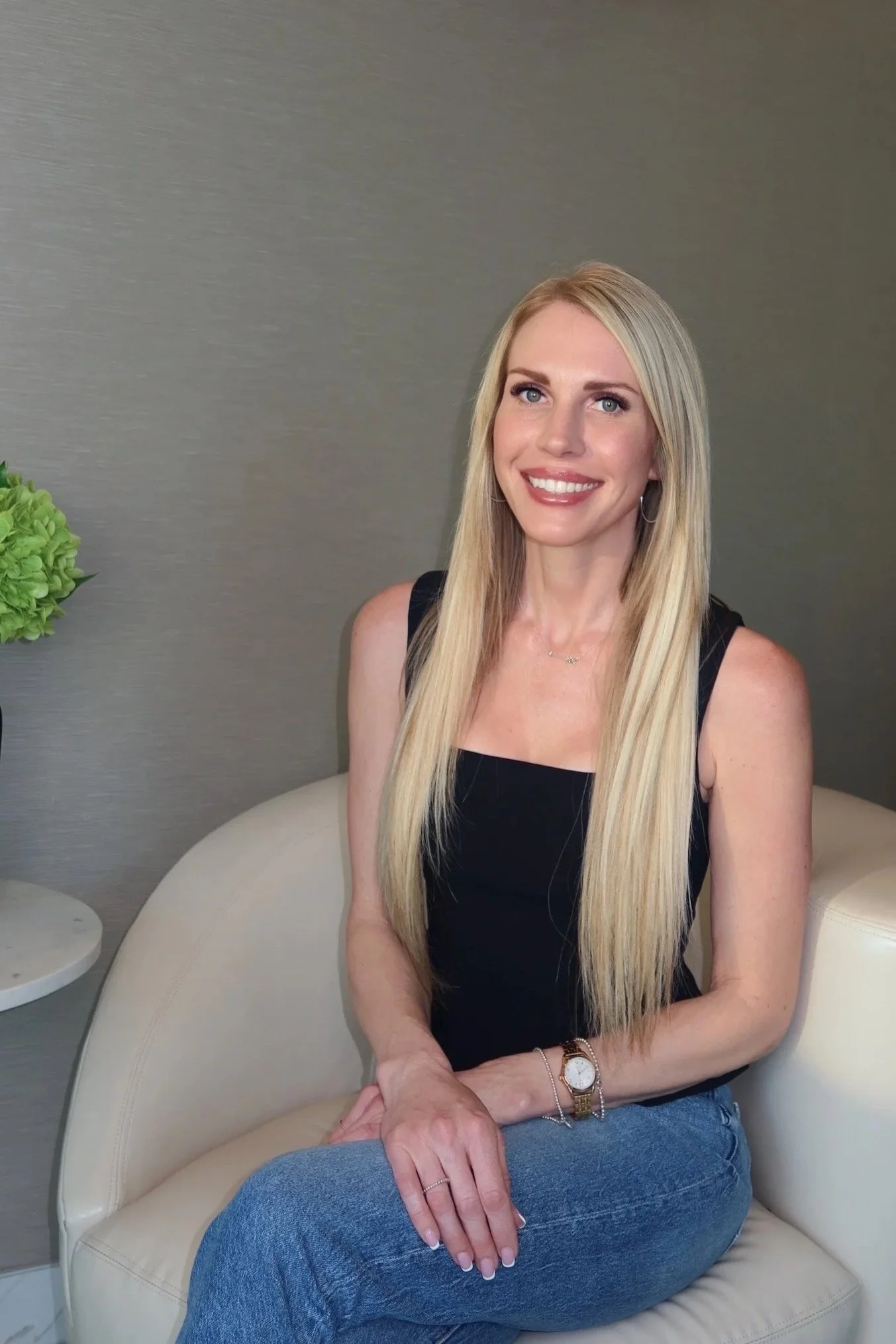 A woman with long blonde hair, blue eyes, and a bright smile, sitting on a beige curved chair in a room with a gray wall, a small white side table, and a green flower arrangement. At the Lift Collective Med Spa in Scottsdale.