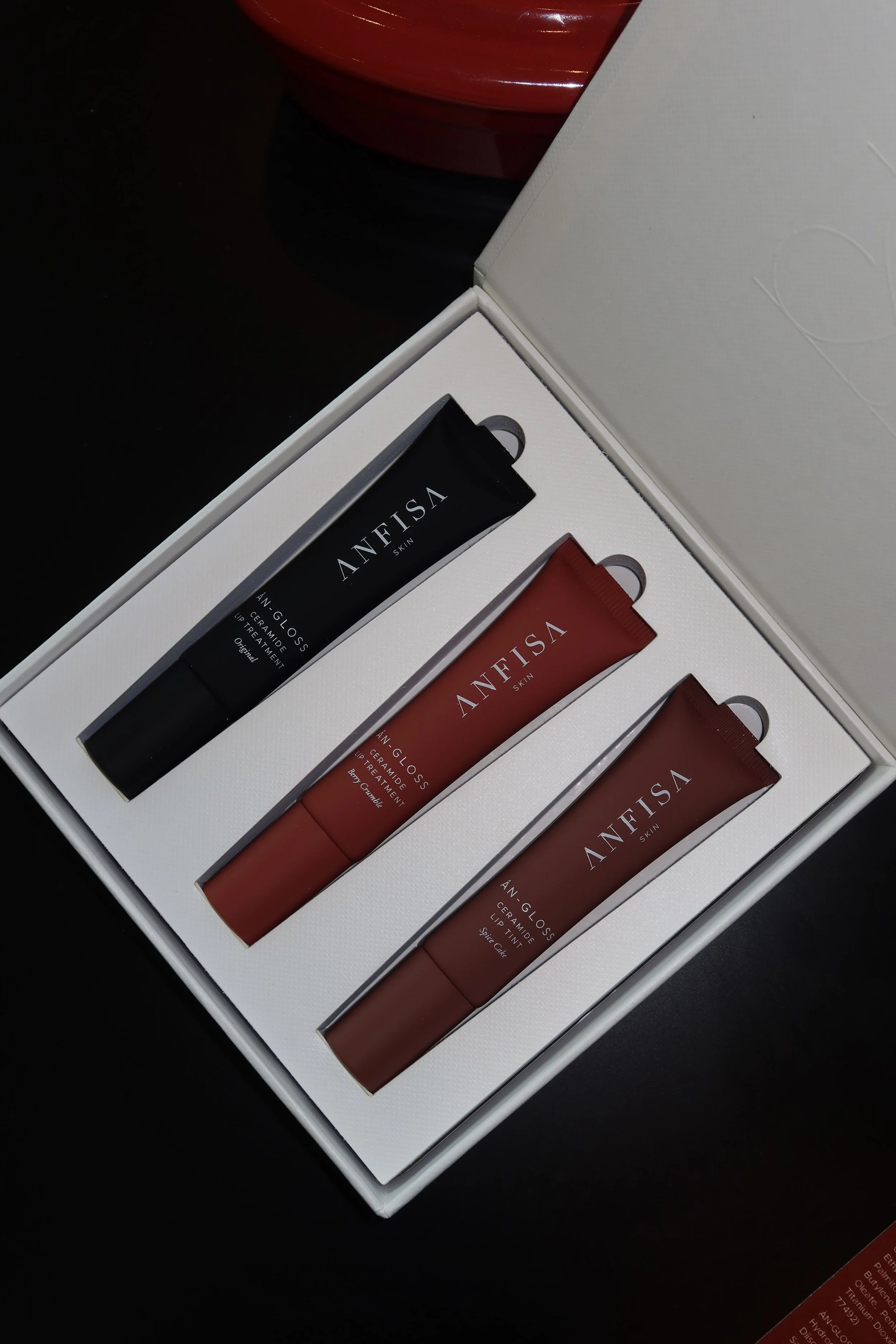 Open white box containing three tubes of ANFISA skincare products, two red and one black, on a black surface with a red container partially visible in the background.