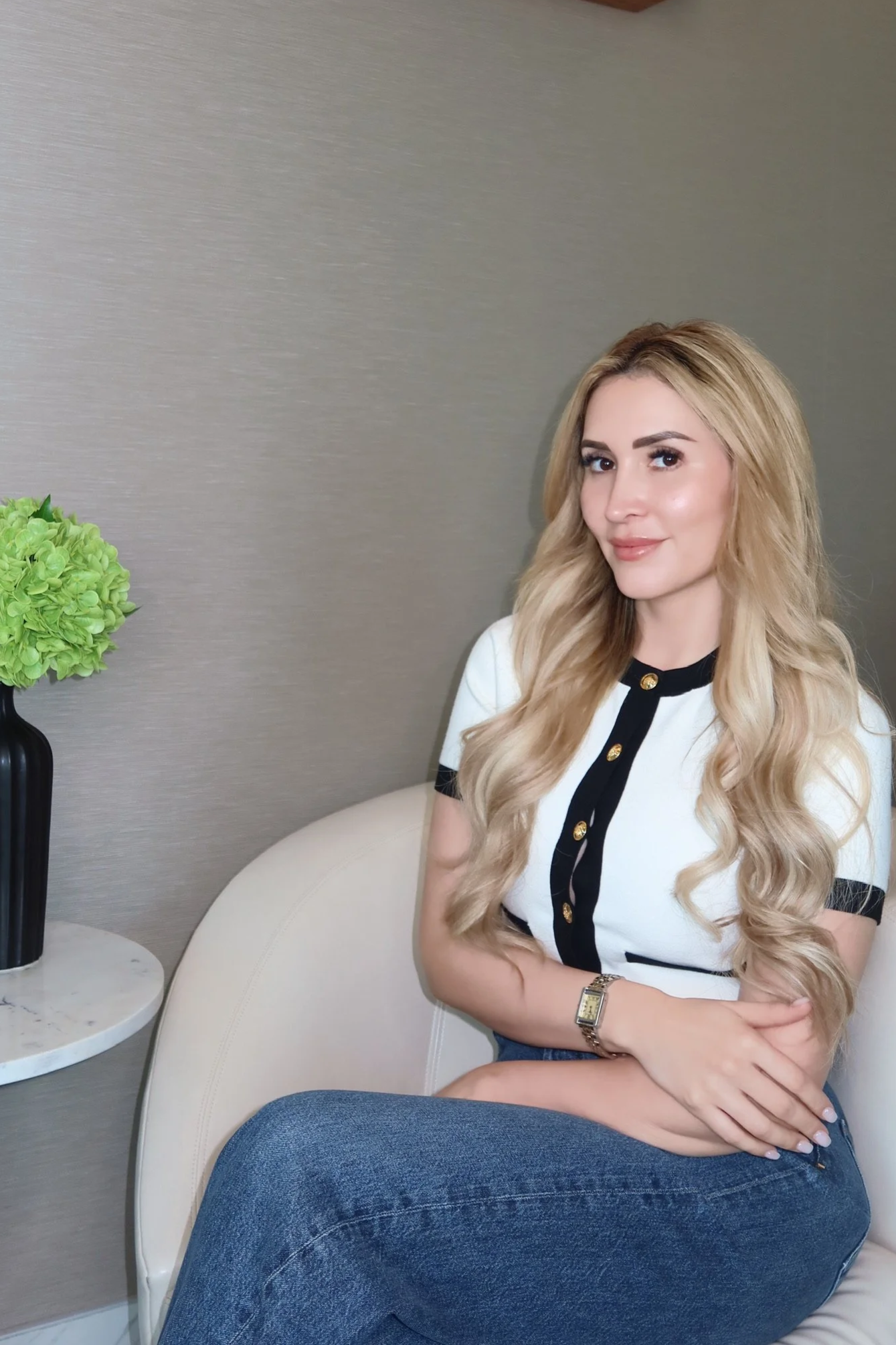 A woman with long blonde hair sitting on a beige couch in a room with a gray wall. She is wearing a black and white top with gold buttons and a gold watch. To her left, there's a black vase with a green hydrangea flower on a small marble side table.