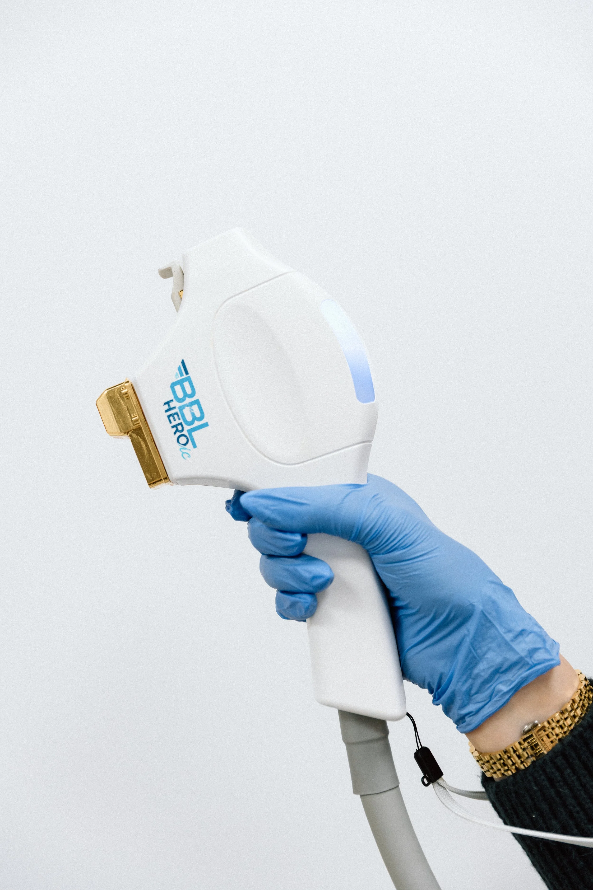 A hand wearing a blue medical glove holding a white laser hair removal device with a gold treatment window, labeled 'BL Herologic,' against a plain white background.