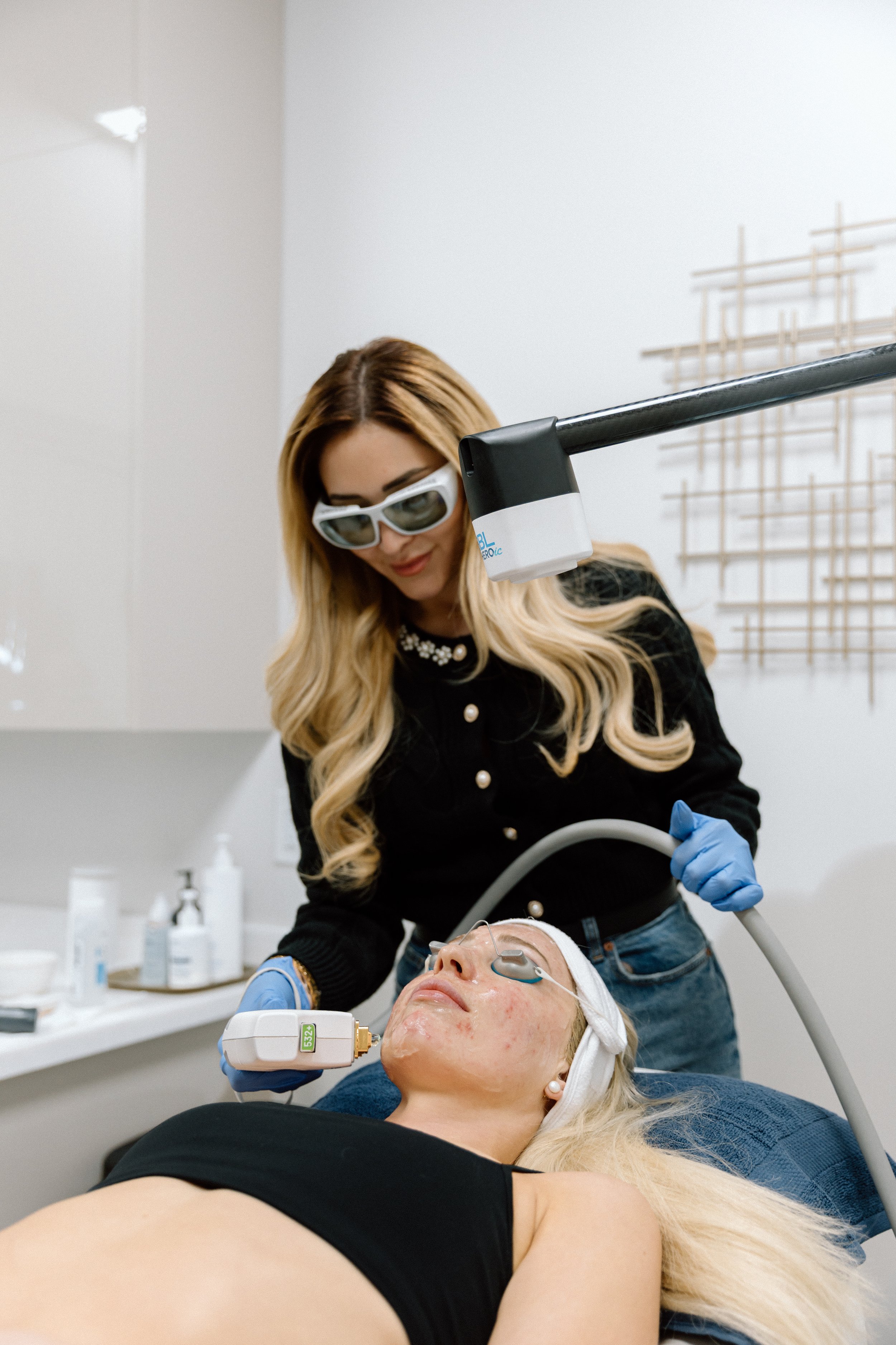 A woman is receiving a laser treatment on her face at a clinic, while a technician wearing protective glasses and gloves operates a large laser device to her face.