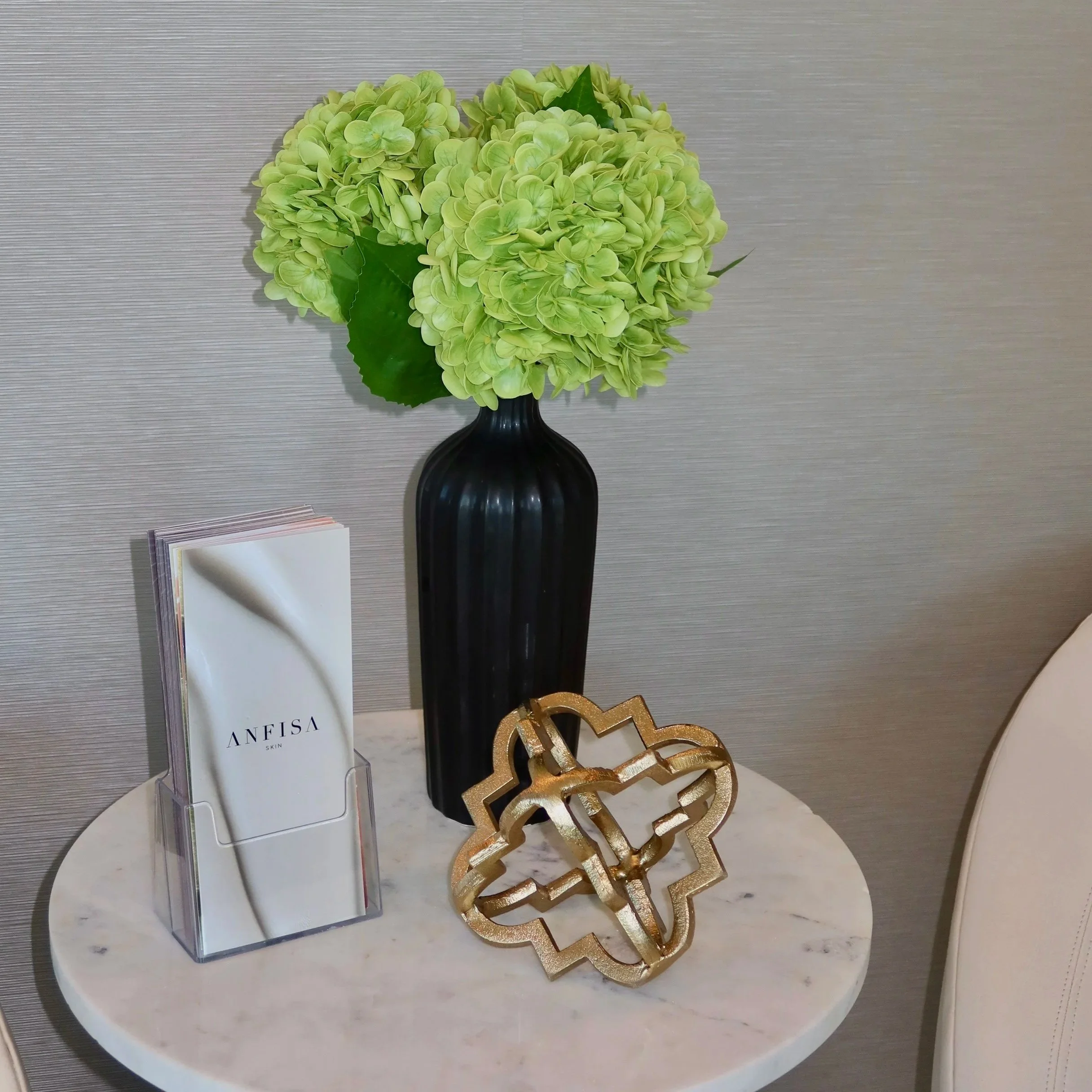 A decorative display featuring green hydrangea flowers in a black vase, placed on a table with magazines and a gold-colored decorative piece. The background includes a textured gray wall. Depicts a luxury med spa.