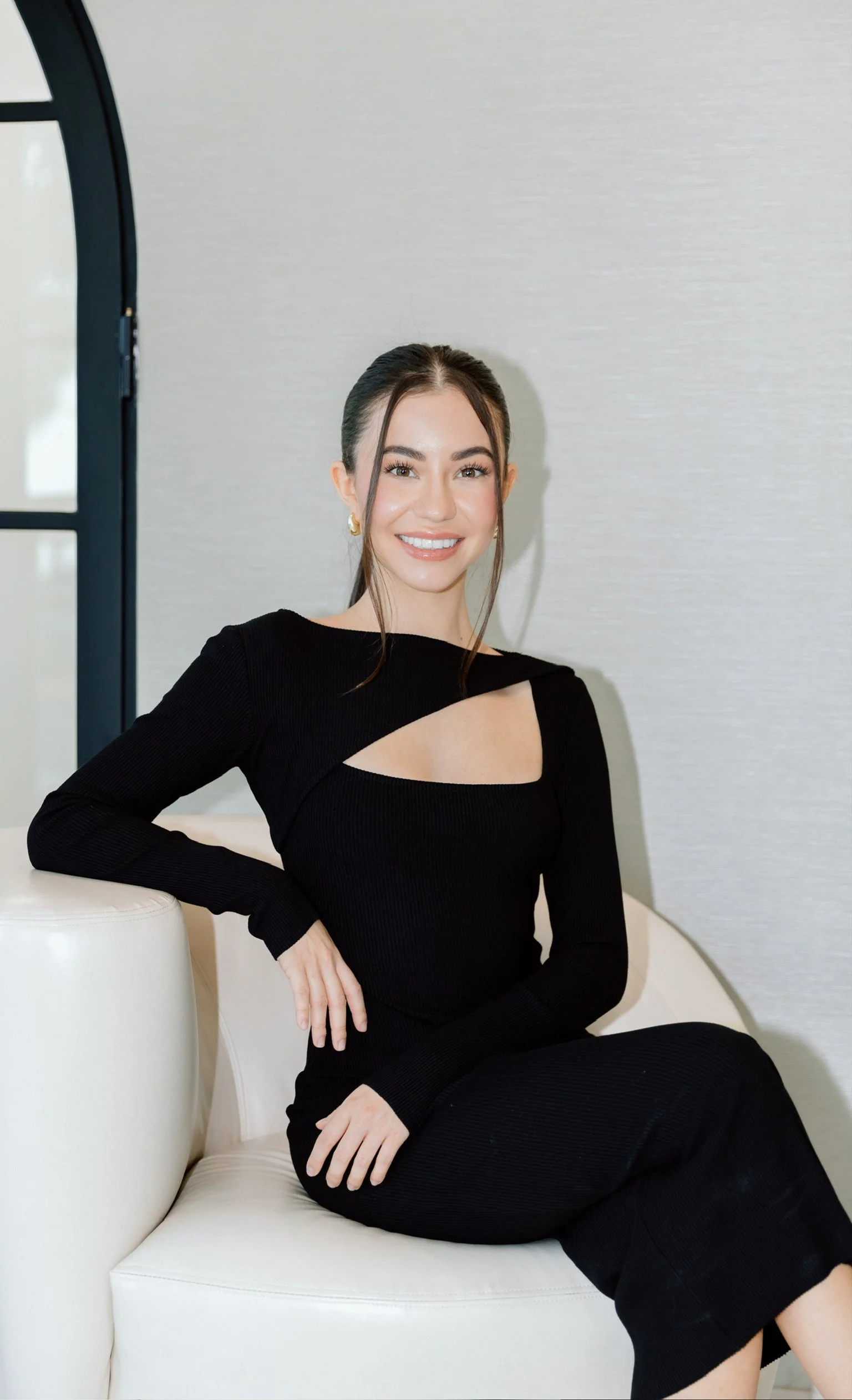 A young woman with dark hair tied back, smiling, wearing a black dress with cut-outs, sitting on a white curved sofa in a room with a grey wall and black window frame.