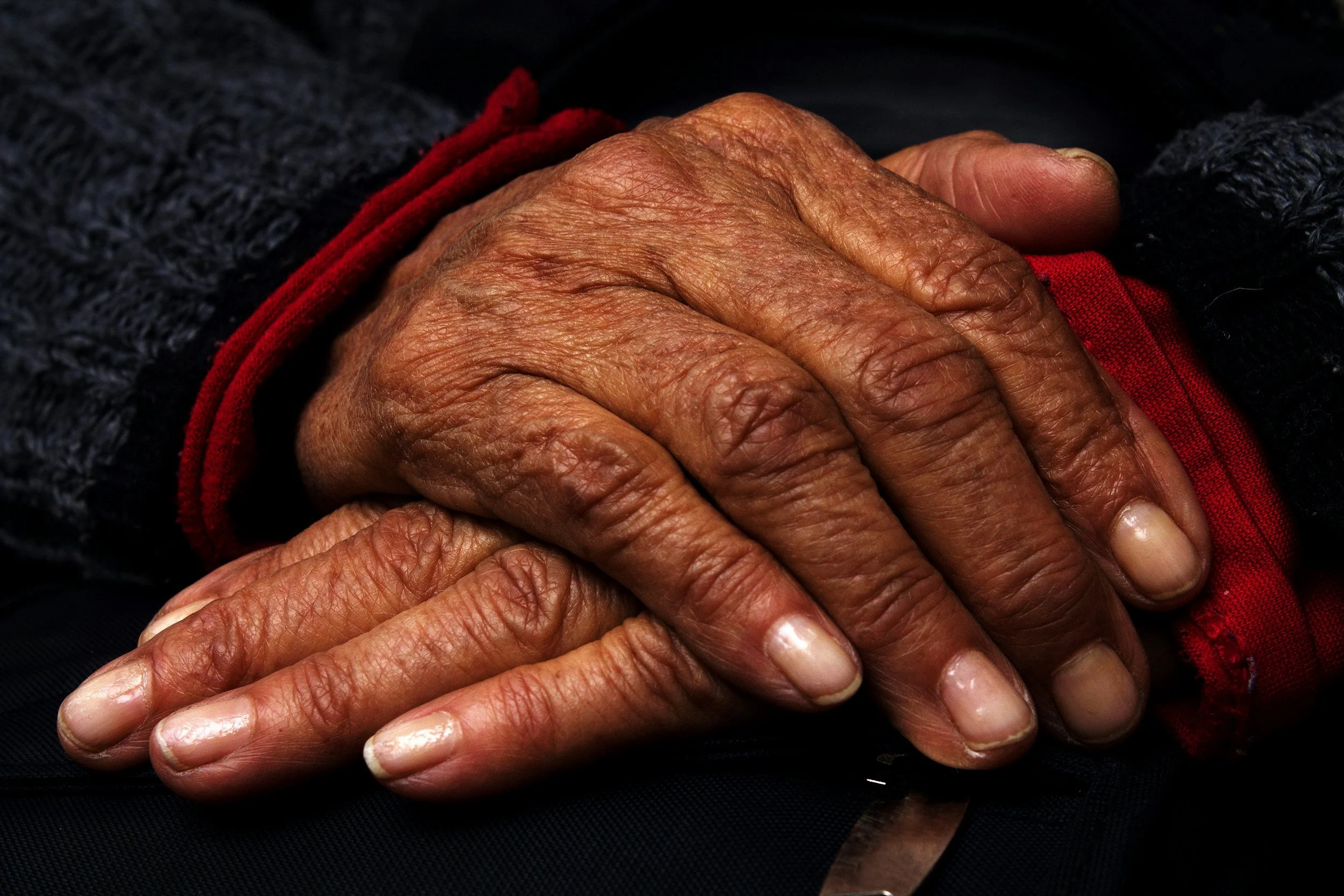 Close-up of two elderly hands clasped together, showing wrinkled skin and nails, resting on dark fabric.