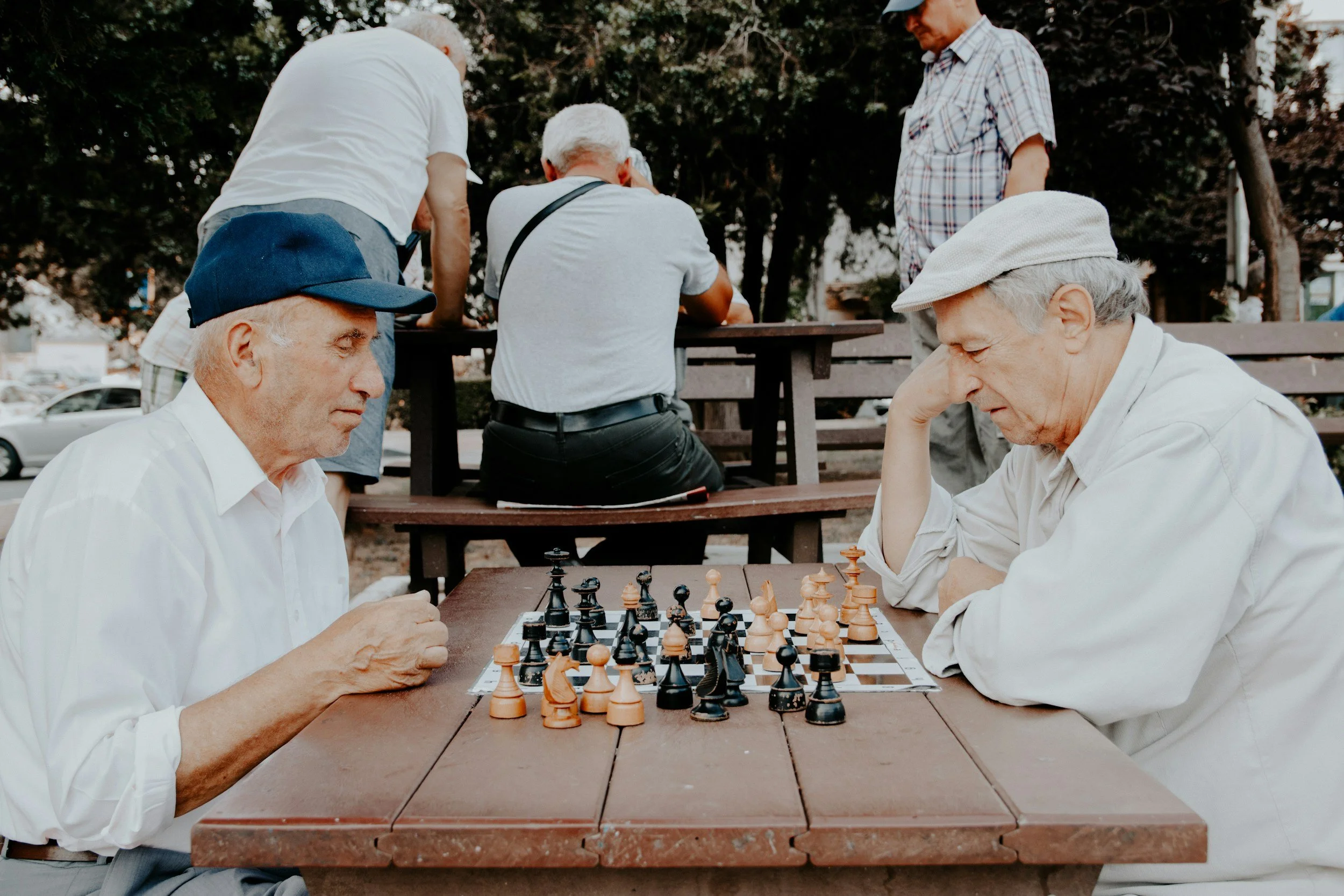 Two elderly men playing chess outdoors at a park, with other people sitting and standing nearby.