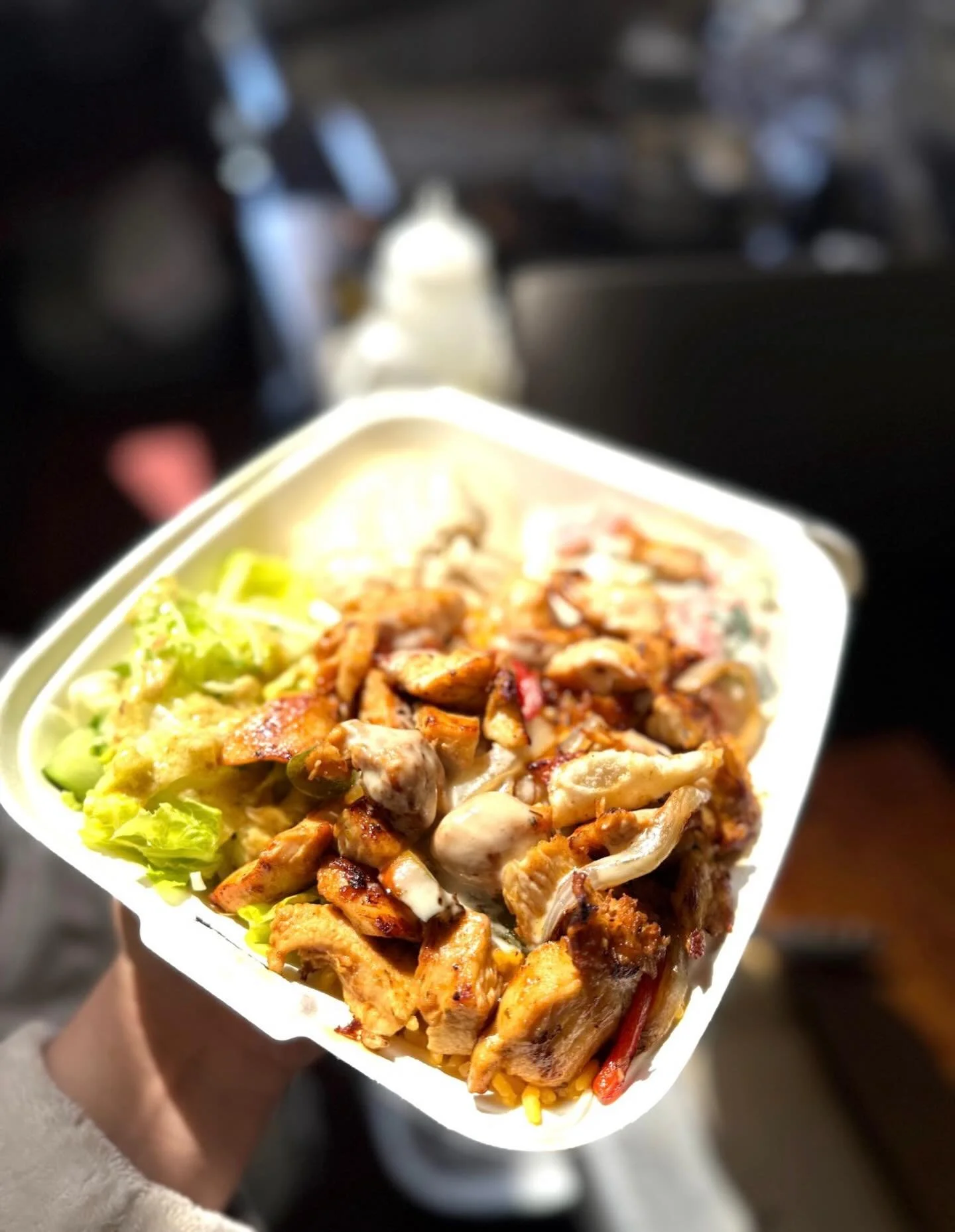 Close-up of a takeout container filled with grilled chicken, vegetables, and rice, held by a person in a moving vehicle.