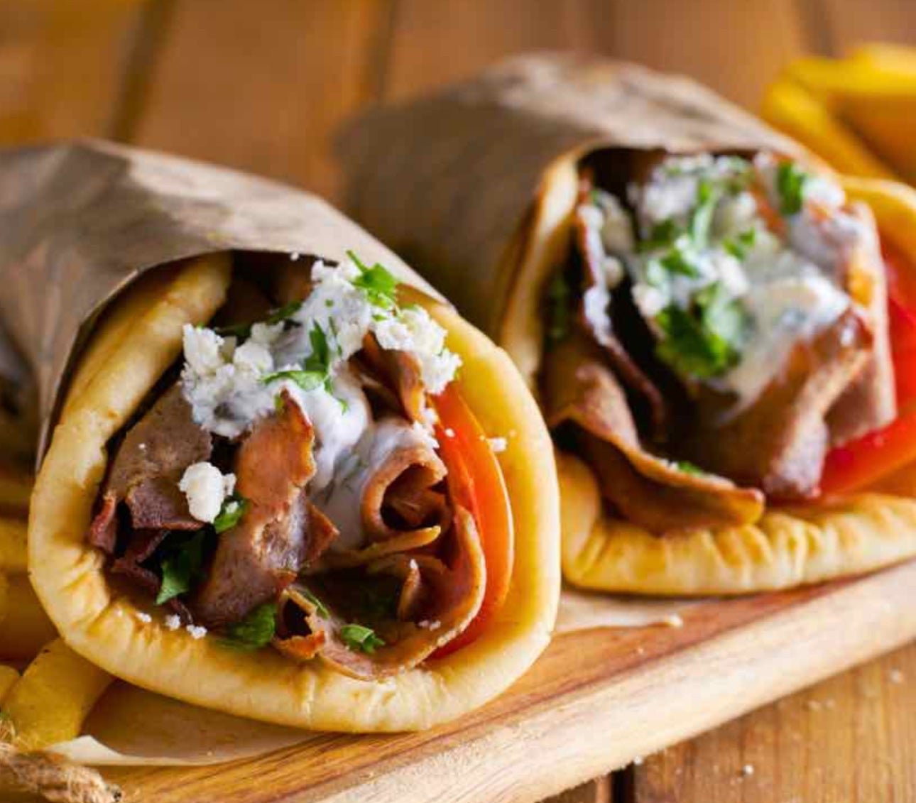 Two burritos filled with beef, tomato, shredded cheese, and cilantro, wrapped in foil, on a wooden serving board.