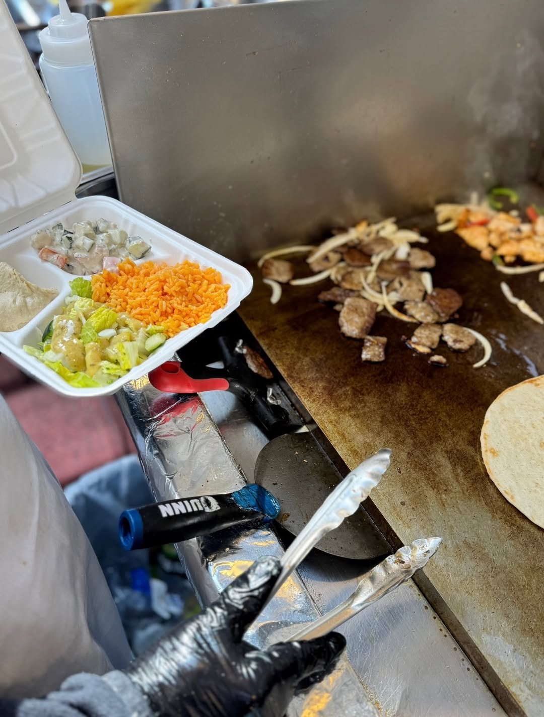 Cook cooking meat, onions, and vegetables on a flat griddle with a large tortilla nearby, while holding a tray of prepared toppings including shredded carrots, diced cucumbers, lettuce, a creamy sauce, and a scoop of hummus.