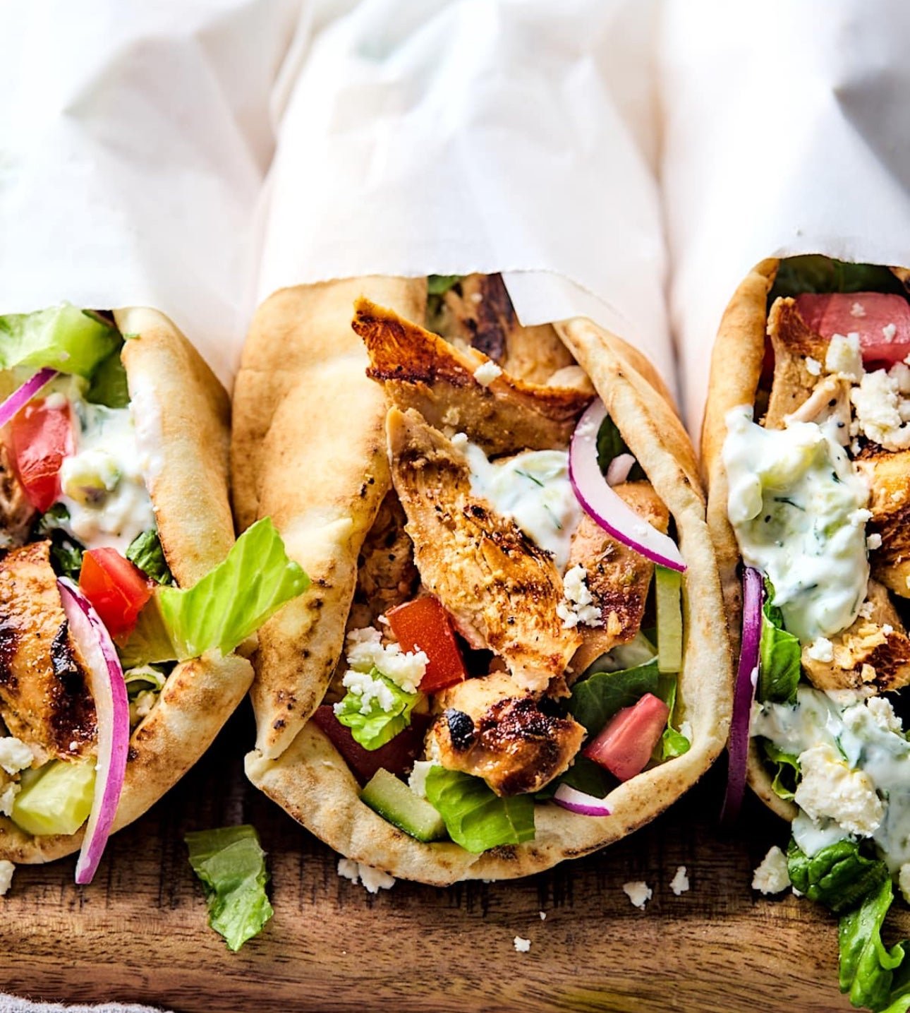 Three chicken gyros sandwiches on flatbread with lettuce, tomatoes, red onions, feta cheese, and tzatziki sauce on a wooden surface.