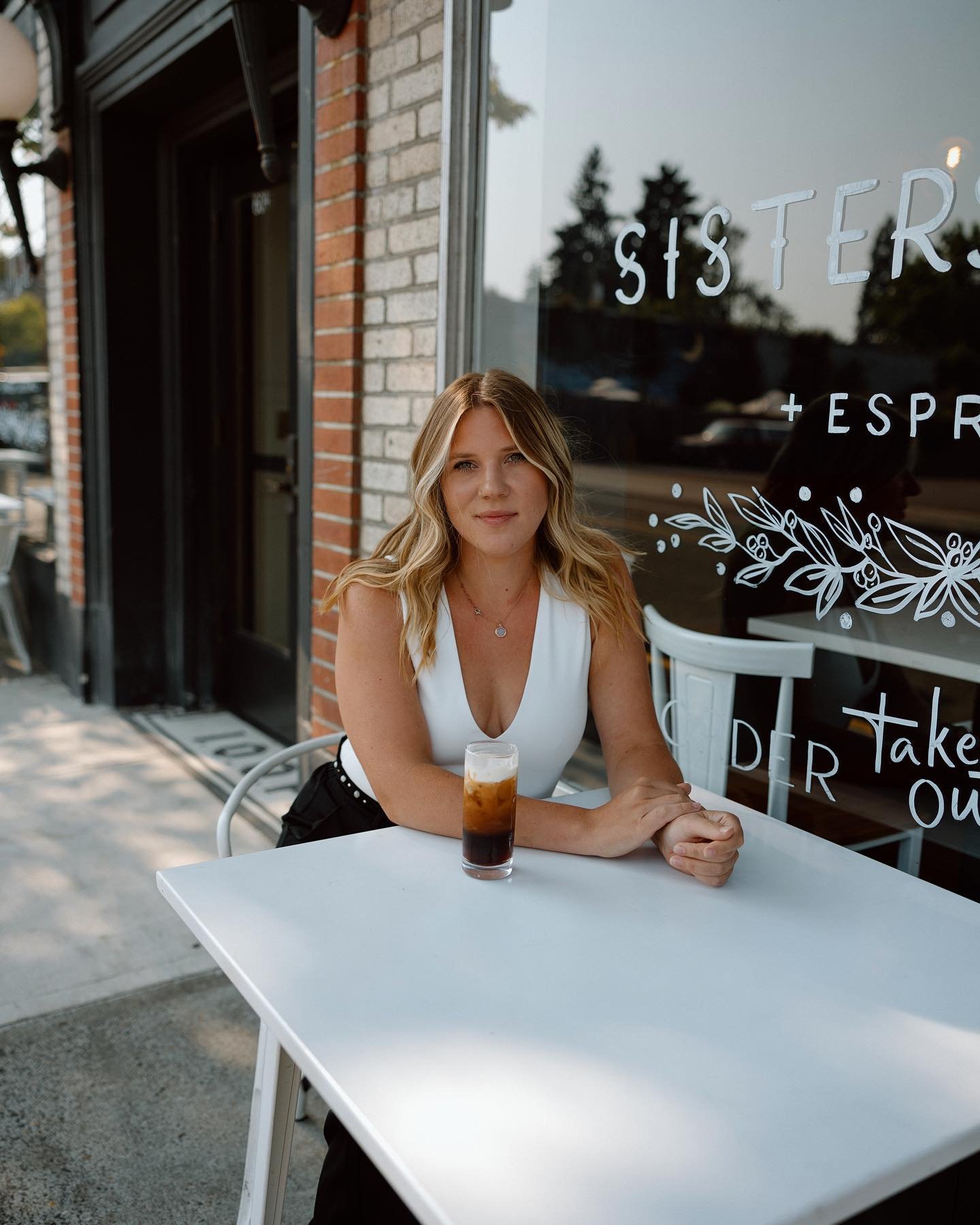 A young woman with blonde hair sitting at an outdoor table of a cafe, with a glass of iced coffee in front of her, in front of a window with the words "SISTERS" and "TAKE OUT" written on it.