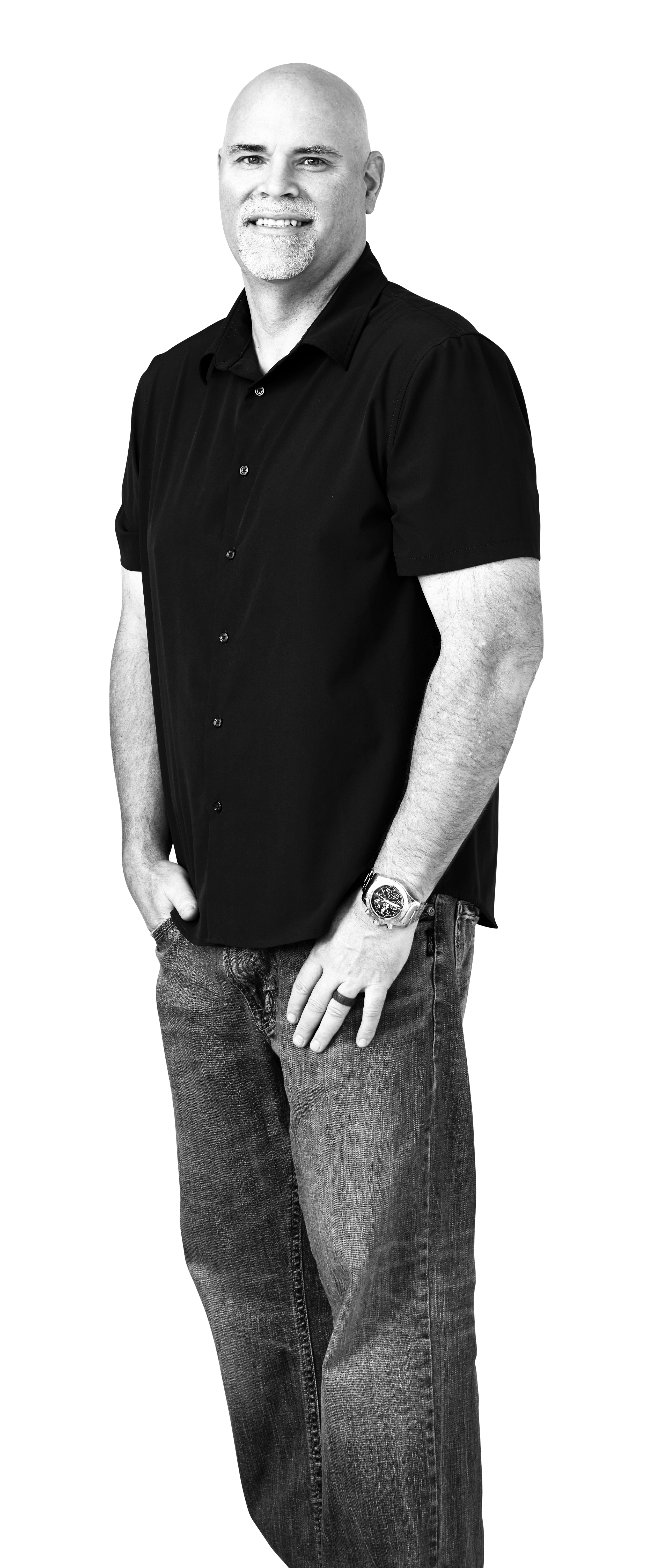 Black and white photo of a bald man with a goatee, wearing a black short-sleeved button-up shirt, jeans, a watch, and a ring, standing with his right hand in his pocket and smiling.
