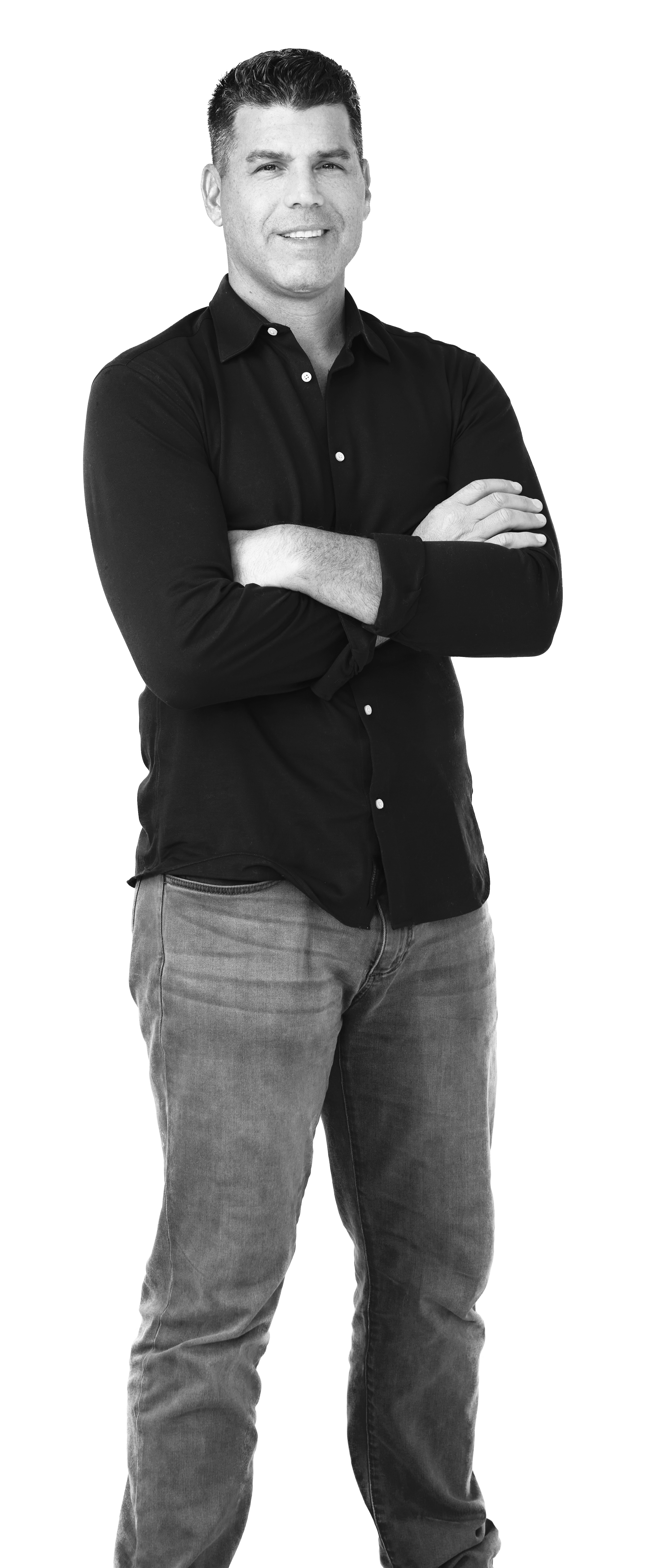 Black and white photo of a man standing with arms crossed, wearing a dark button-up shirt and jeans, smiling.