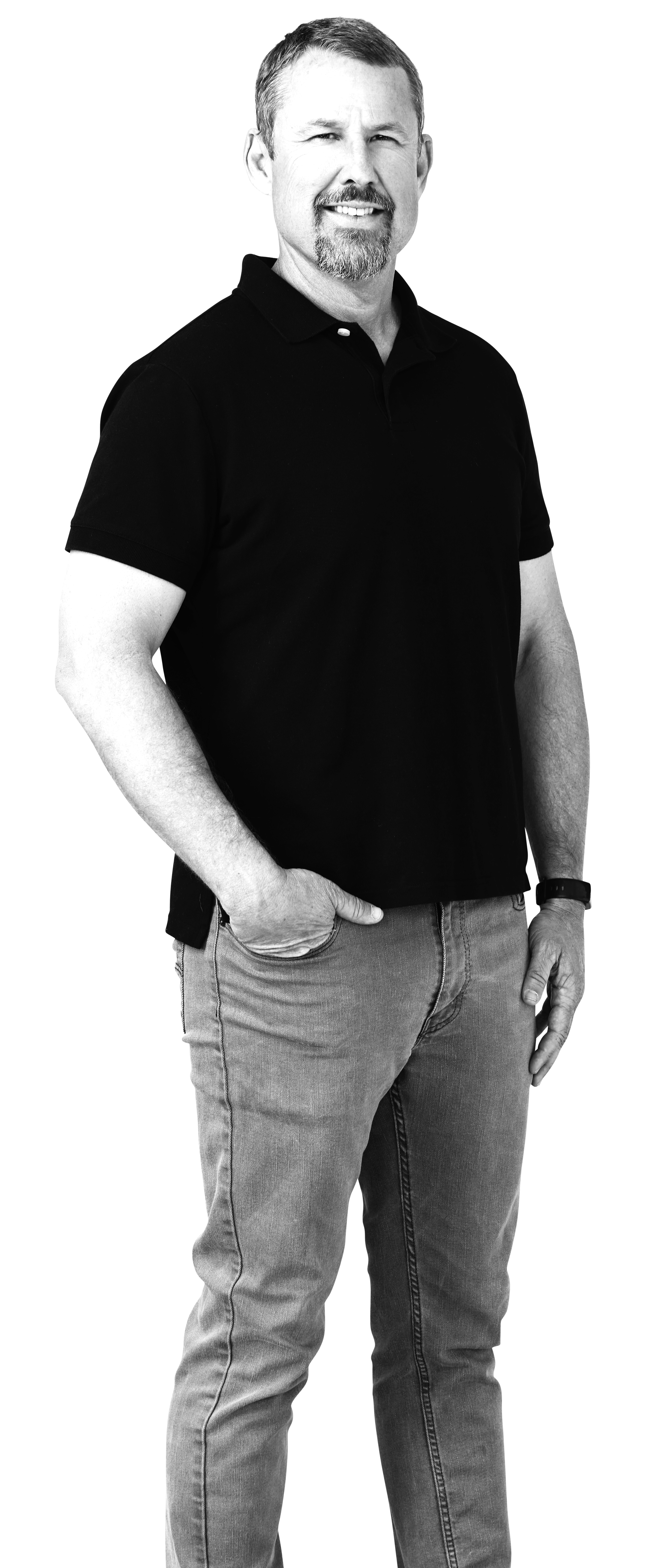 A man with short hair, a goatee, and a mustache standing against a neutral background. He is wearing a black polo shirt, jeans, and a watch, with one hand in his pocket and the other at his side, smiling slightly.