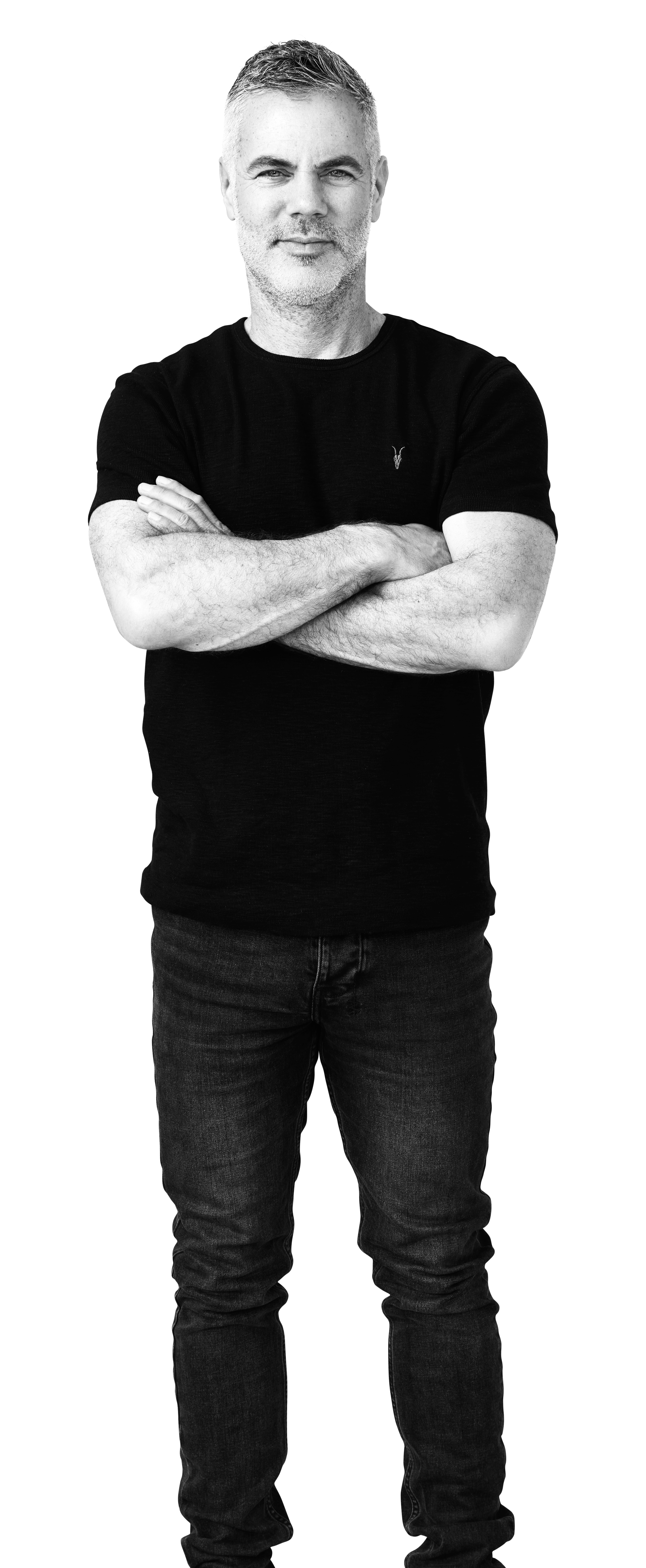 Black and white photo of a middle-aged man standing with arms crossed, wearing a black t-shirt and jeans, smiling slightly.