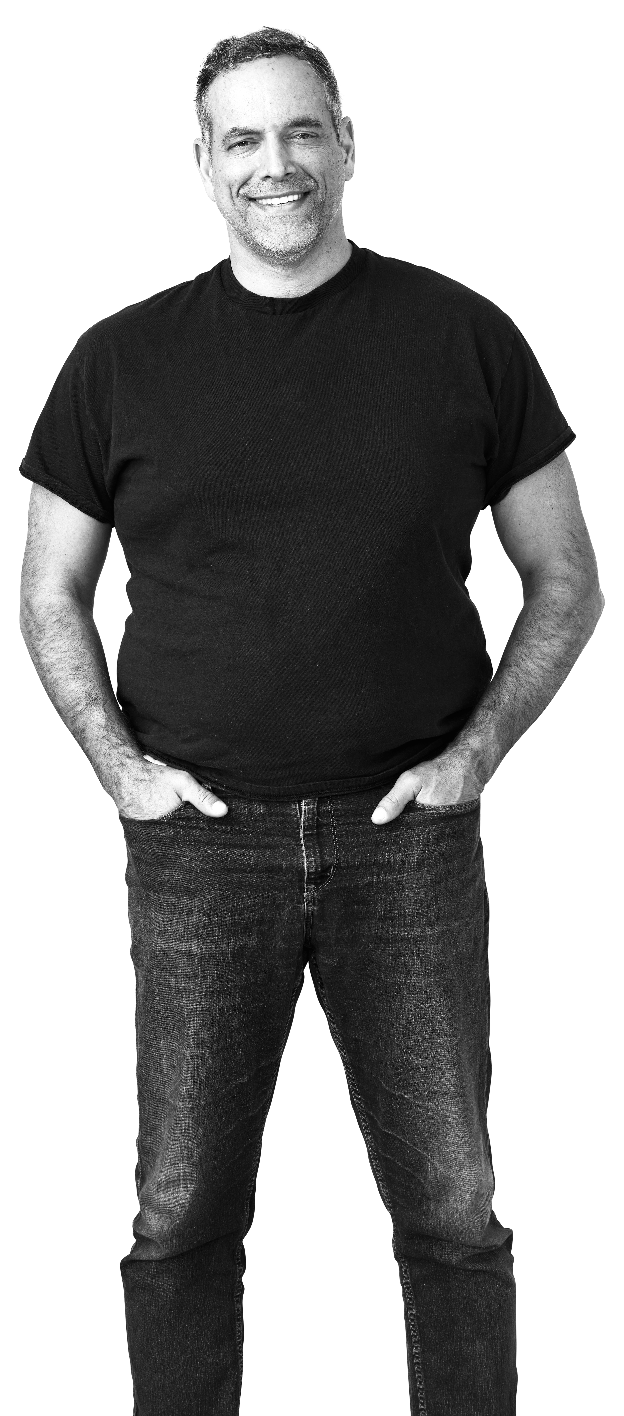 A smiling man with short dark hair, a beard, dressed in a black t-shirt and jeans, standing with hands in pockets against a dark background.