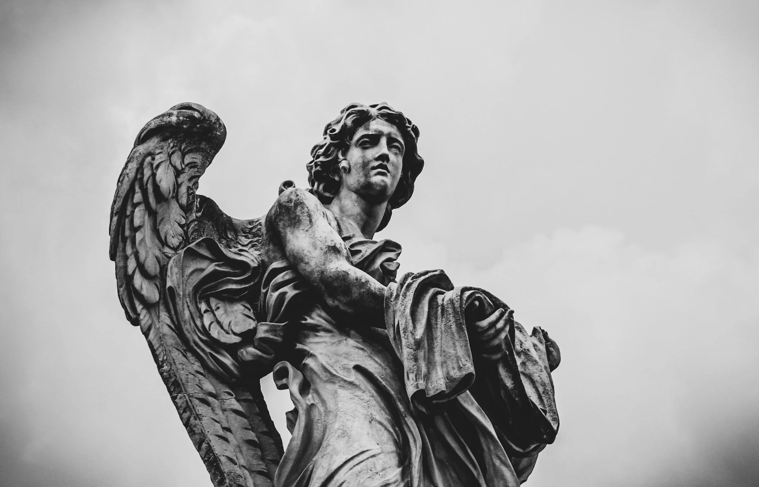 Black and white photograph of a detailed stone statue of an angel with long, curly hair and large wings, looking off into the distance against a cloudy sky.