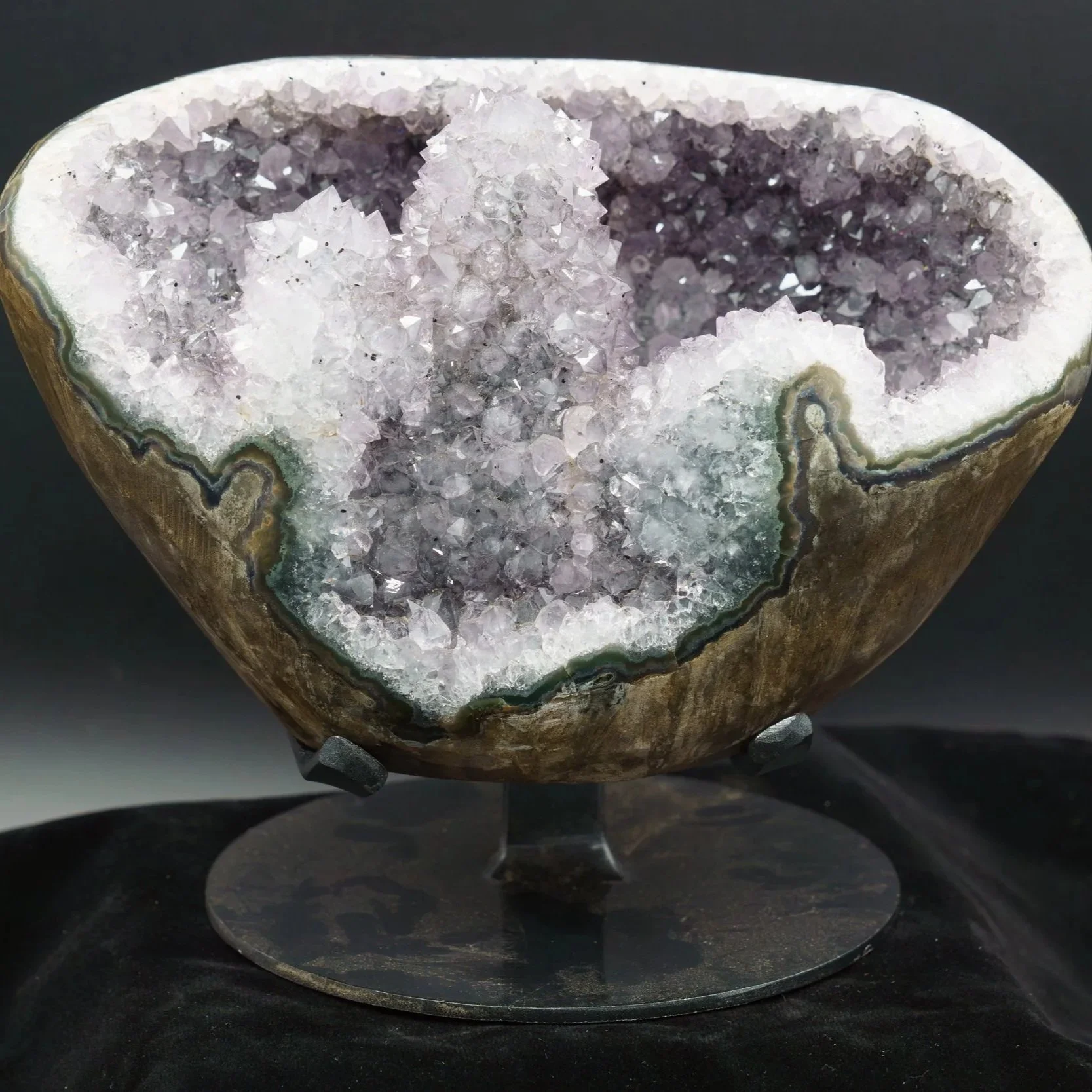 A polished geode with a hollow interior filled with purple and clear crystals, mounted on a stand against a black background.