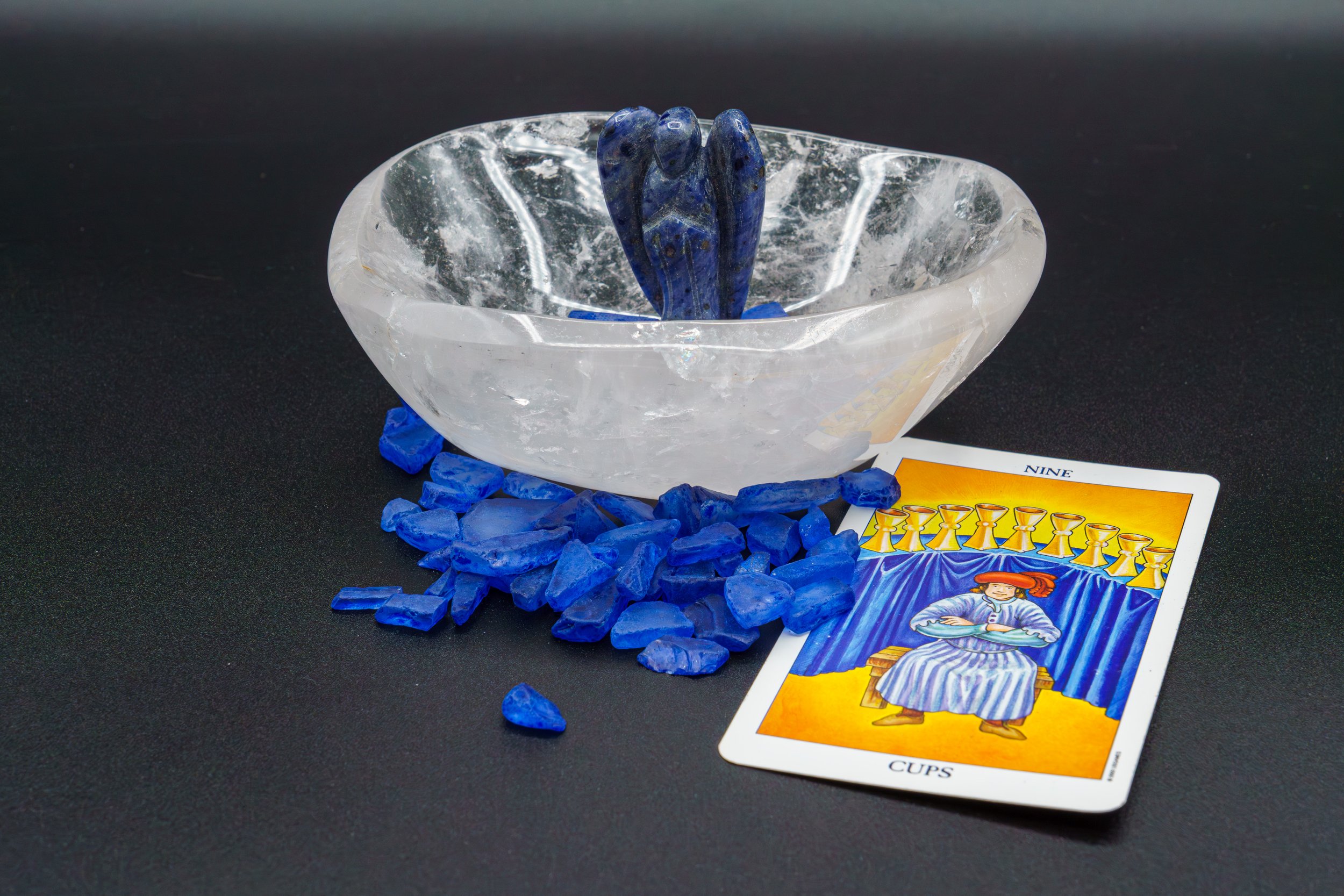 A crystal bowl with a blue gemstone carved into the shape of praying hands inside, surrounded by scattered blue stones and tarot cards showing the Nine of Cups.