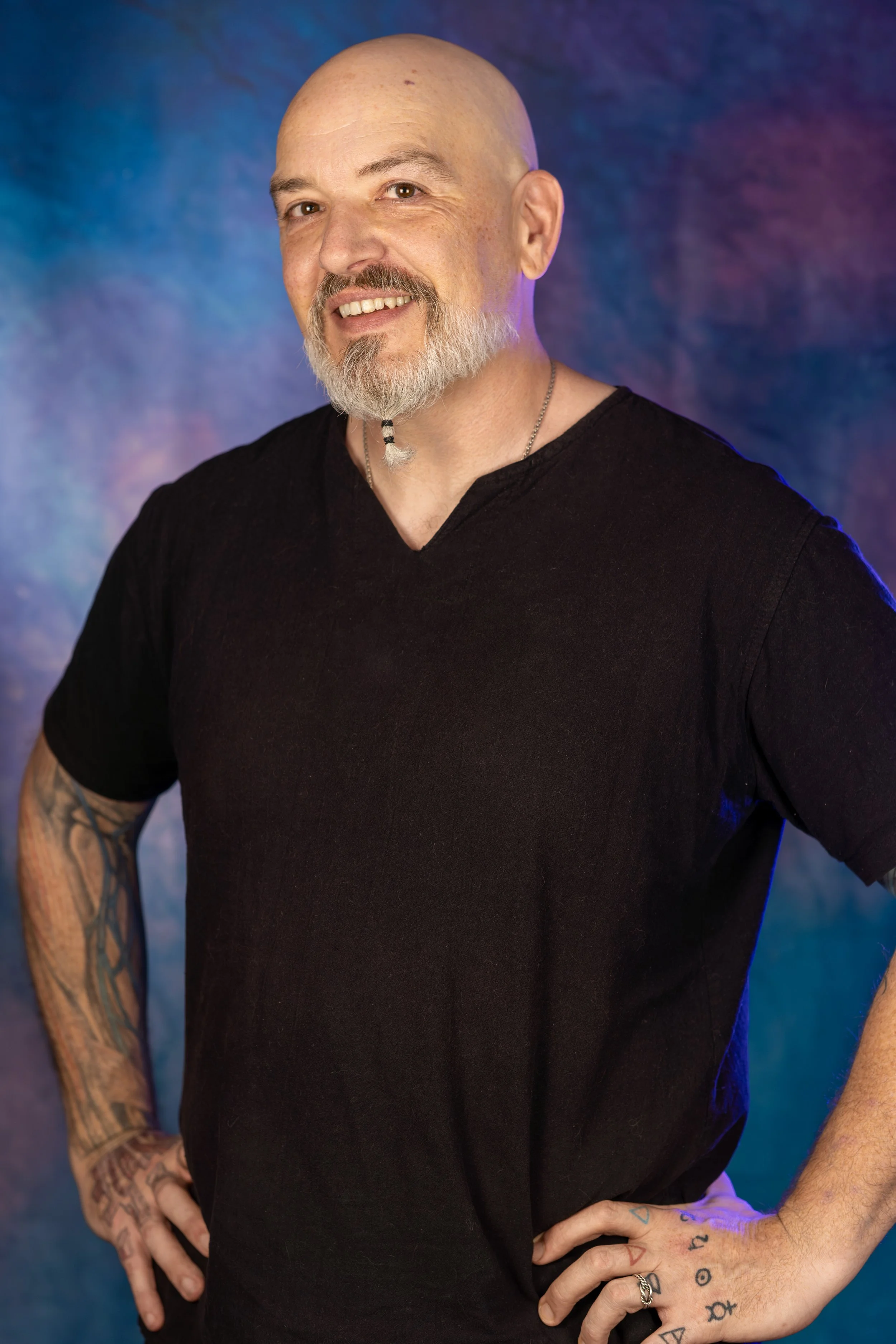 A middle-aged man with a shaved head, gray beard, and tattoos on his arms, smiling and posing with hands on hips in front of a colorful, abstract background.