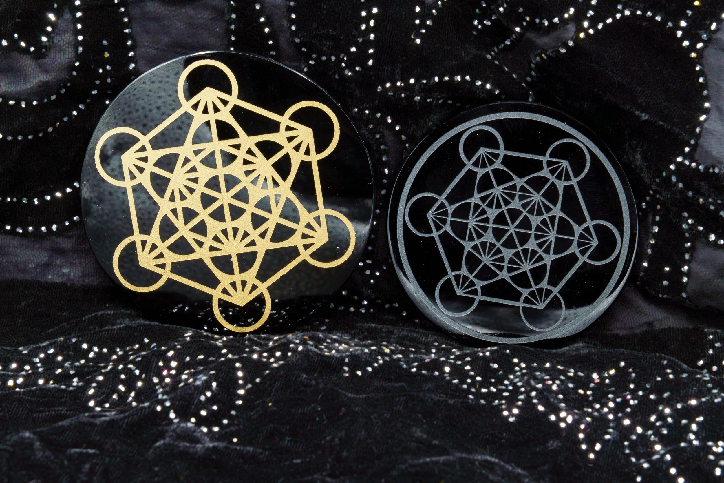 Two round decorative pieces with intricate geometric patterns, one gold and one silver, placed on a black fabric with shiny dotted embellishments.