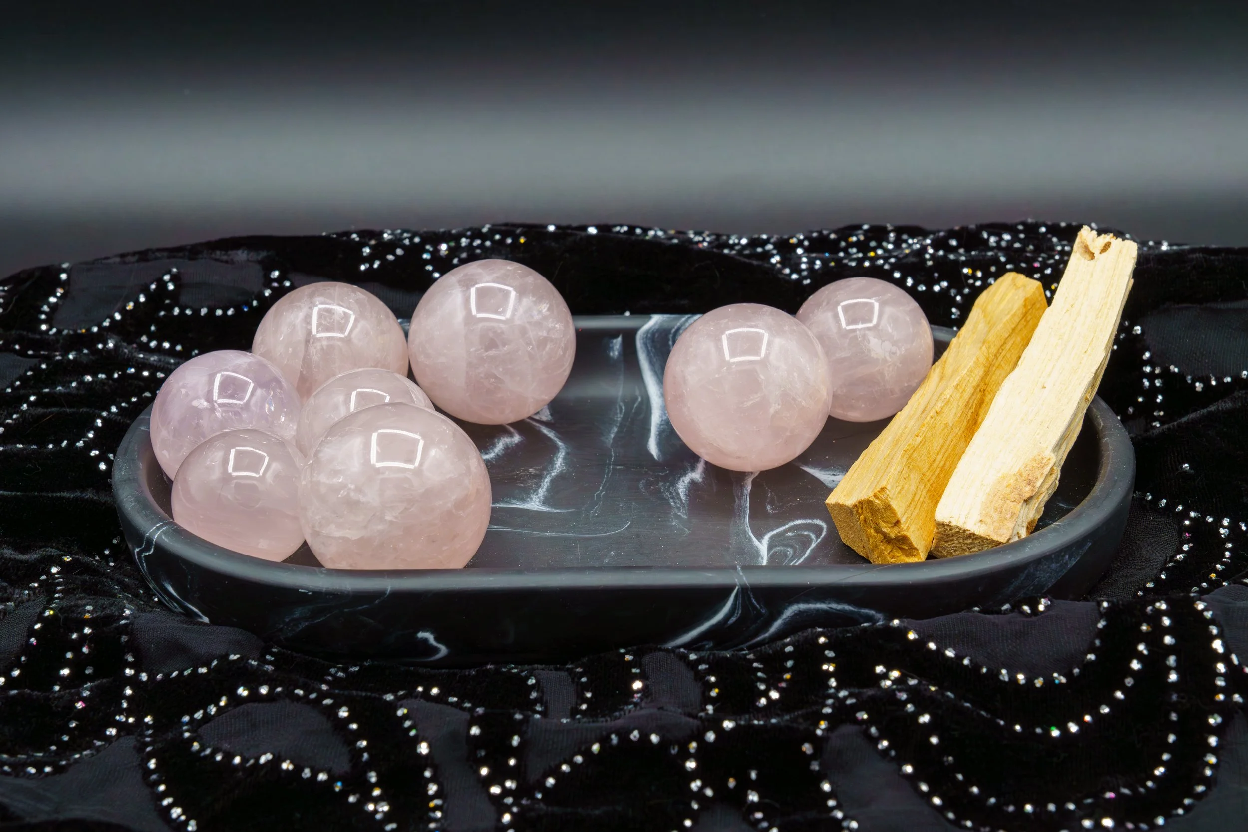 Pink rose quartz spheres and two palo santo wood sticks on a shiny black fabric with glittering details.