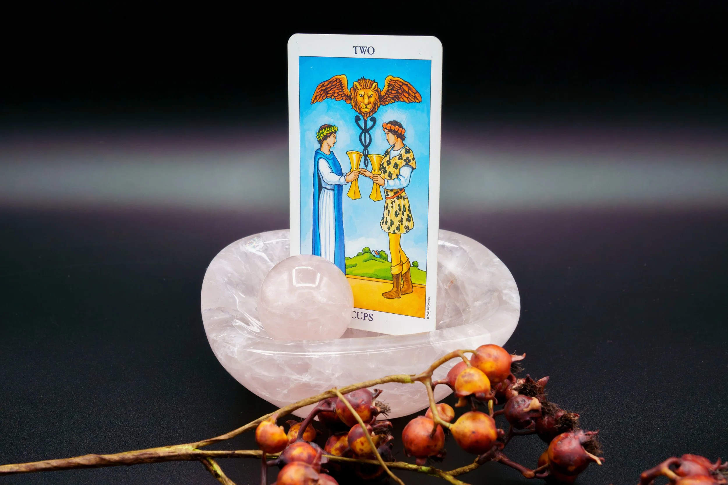 Tarot card of the Two of Cups resting on a large pinkish-white salt crystal, with small red and purple berries on a branch in the foreground.