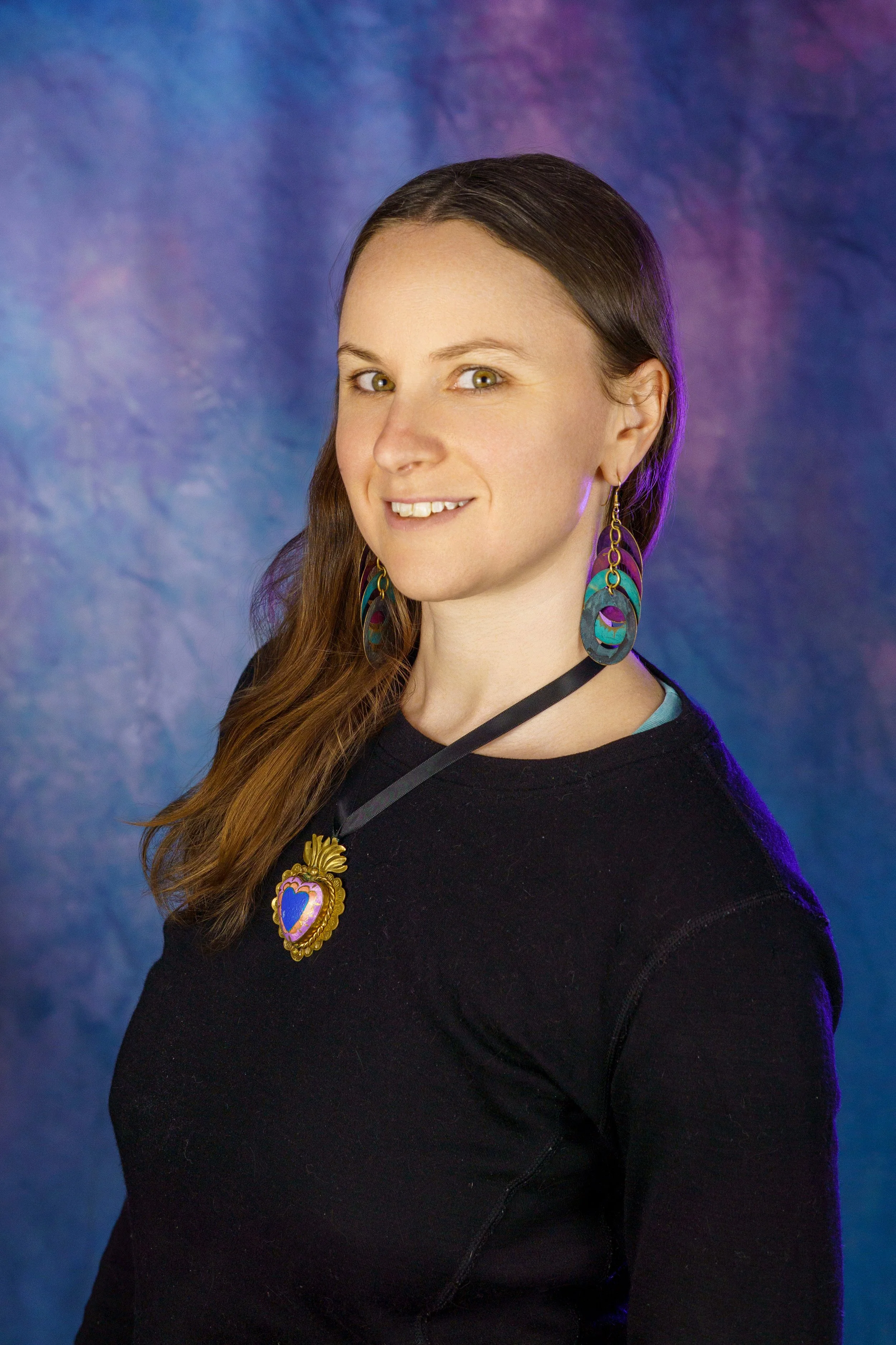 A woman with long brown hair wearing large colorful earrings and a decorative necklace, smiling with a dark backdrop featuring blue and purple hues.
