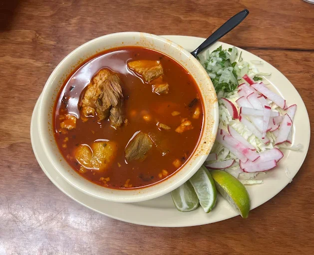 A bowl of beef stew with potatoes in a red broth, served with chopped onions, cilantro, and lime wedges on the side.
