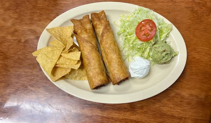 Plate with three taquitos, tortilla chips, lettuce, slice of tomato, scoop of guacamole, and dollop of sour cream.