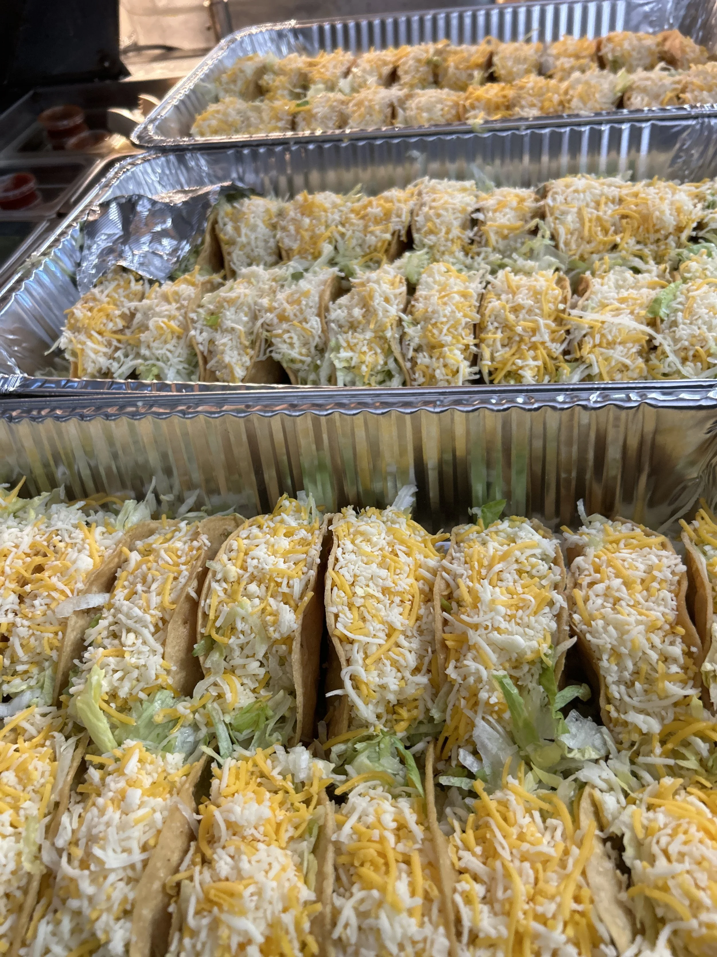Three aluminum trays filled with shredded cheese tacos topped with shredded cheese and lettuce.