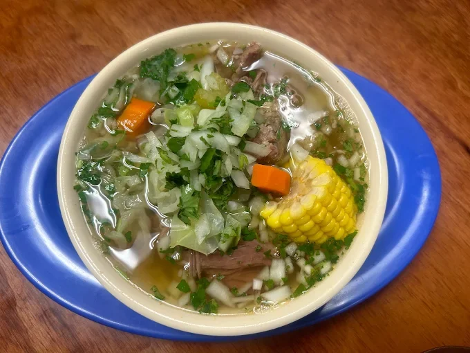 A bowl of beef vegetable soup with corn on the cob, carrots, chopped onions, and herbs, served on a blue plate on a wooden table.