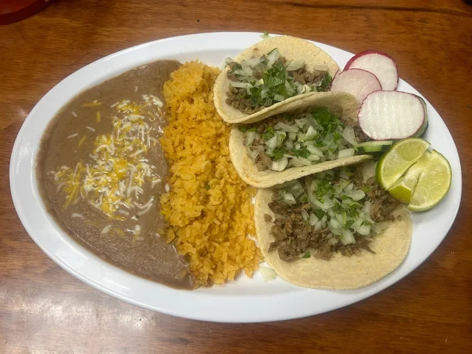 Plate of Mexican food with rice, refried beans topped with cheese, and three tacos filled with seasoned beef and topped with chopped onions and cilantro. Garnished with sliced radishes and lime wedges.