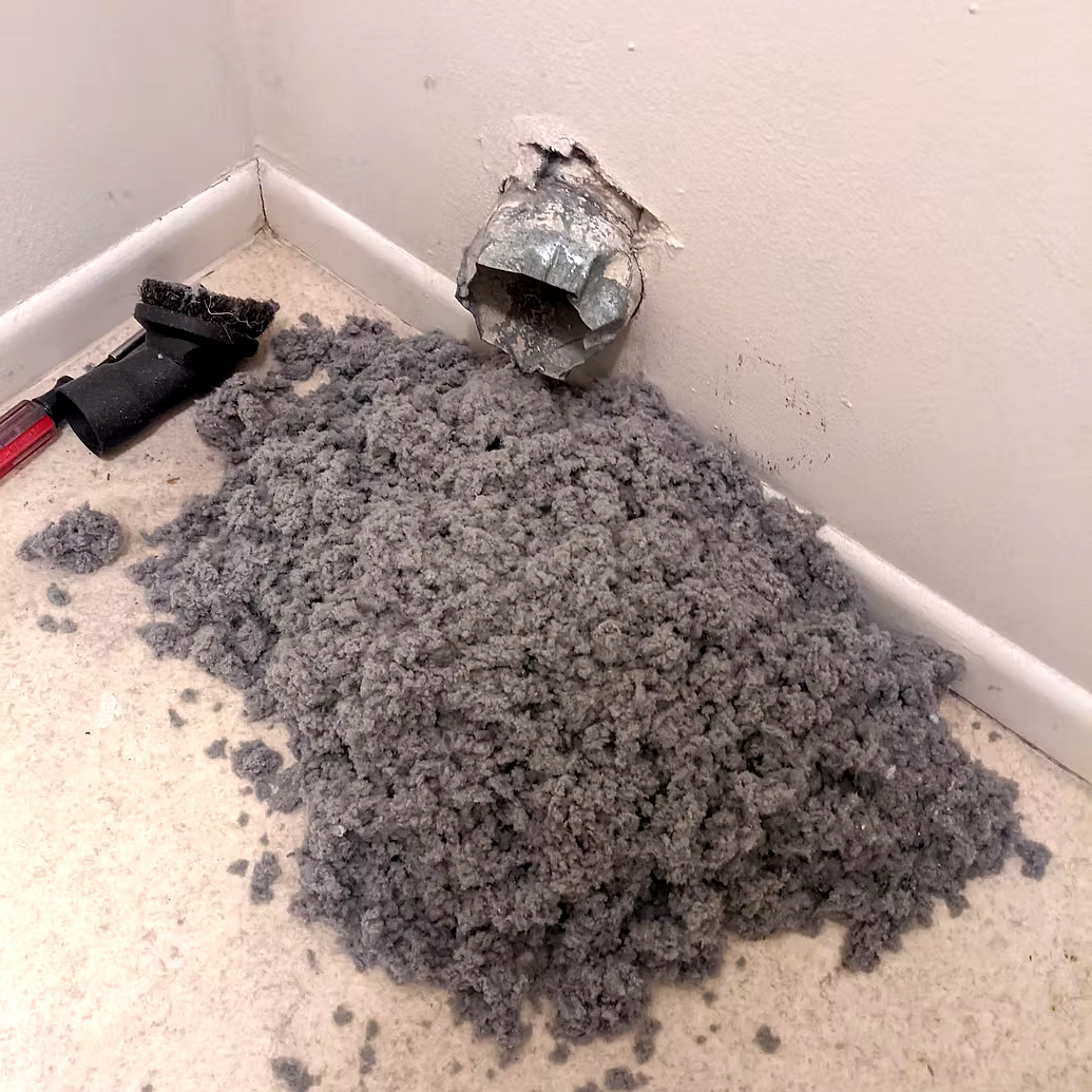 Floor repair with a pile of gray insulation or debris, a floor vent duct, and a utility brush on the floor near a wall corner.