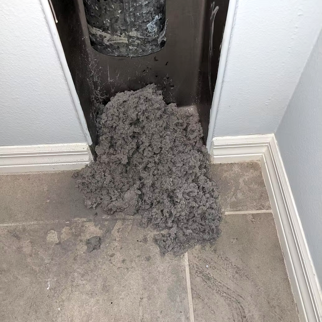 Clump of black insulation foam spilled on gray tiled floor near the base of an appliance or vent.