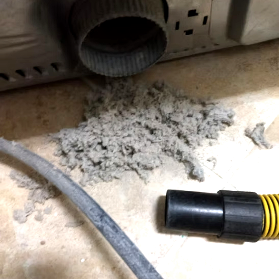 Dust and debris on a floor near a vent or pipe, with a vacuum attachment nearby.