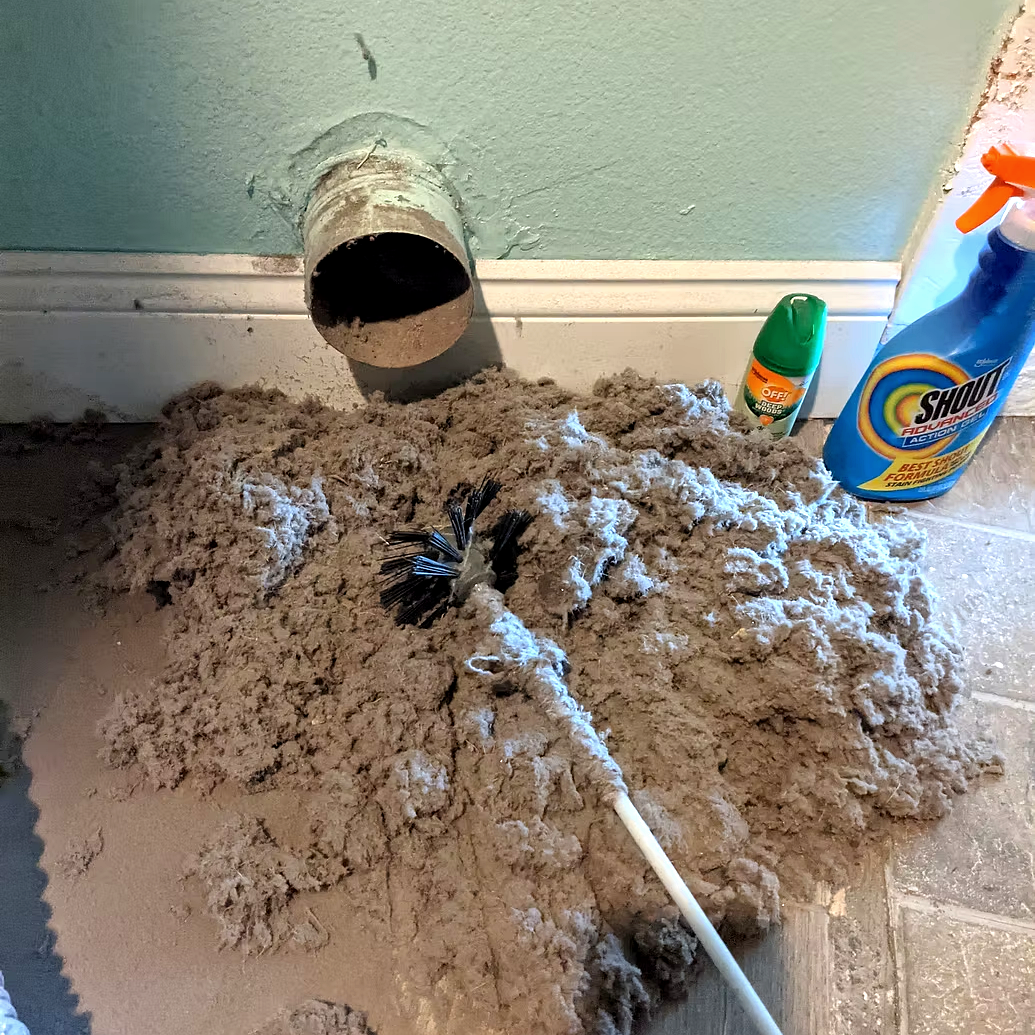 Clogged pipe emitting debris and water, surrounded by cleaning supplies including a brush, spray bottle, and cleaning products on the floor.