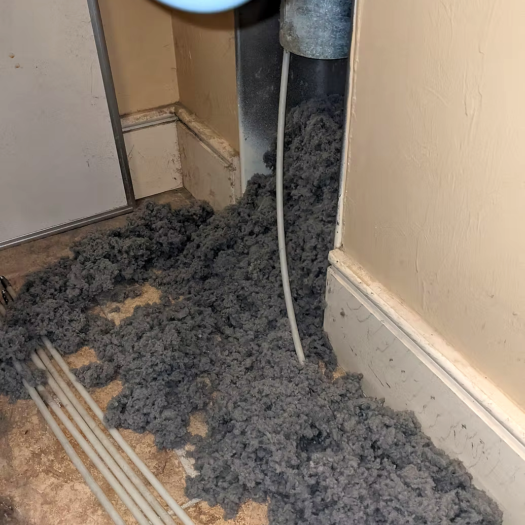 A construction site with gray insulation material spilled on the floor near a wall, electrical wiring, and pipes.