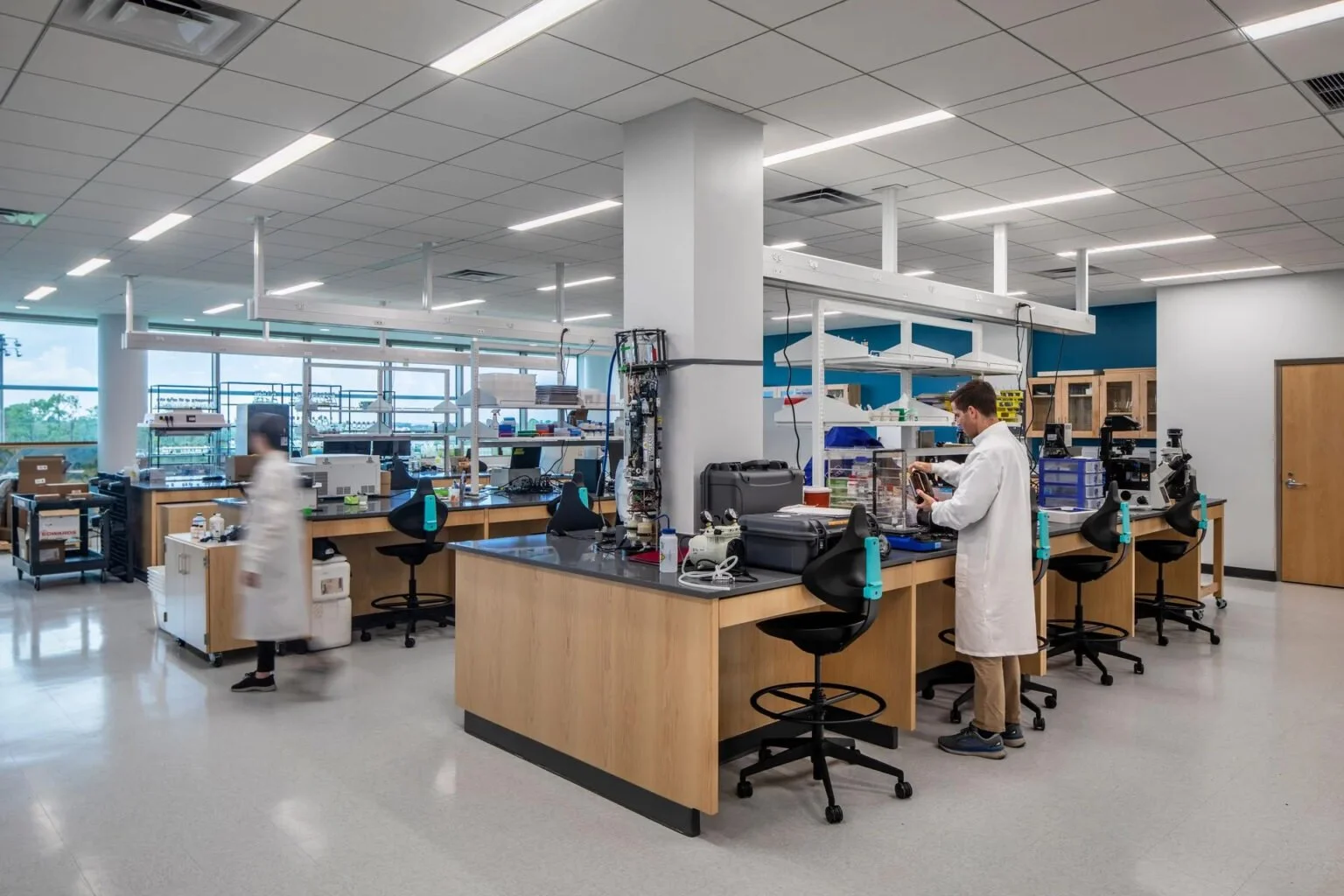 A laboratory with scientists working at lab benches, equipped with scientific instruments, glassware, and supplies. The room has large windows, tiled flooring, and bright, even lighting.